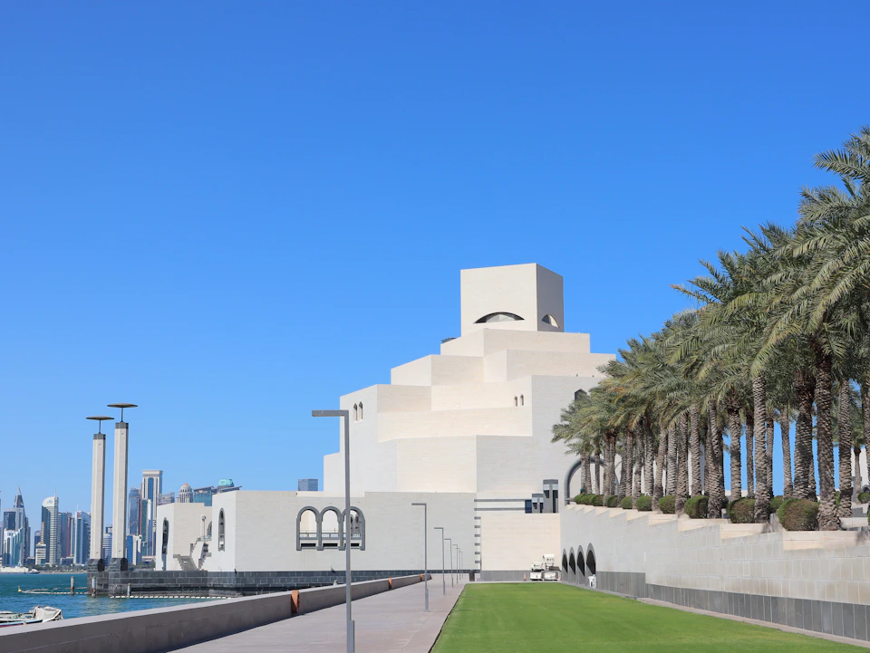 The image shows a large, white building with a distinctive pyramid-like structure on top, situated near the water's edge and surrounded by palm trees. The building is part of the Dubai Museum, which is located within the cityscape of Dubai International Airport. In the foreground, there are several palm trees that add to the tropical ambiance of the scene.

The photograph captures a clear day with a blue sky overhead, providing an ideal backdrop for the white building and palm trees. The perspective of the image offers a comprehensive view of the museum's architectural features and its integration into the cityscape.