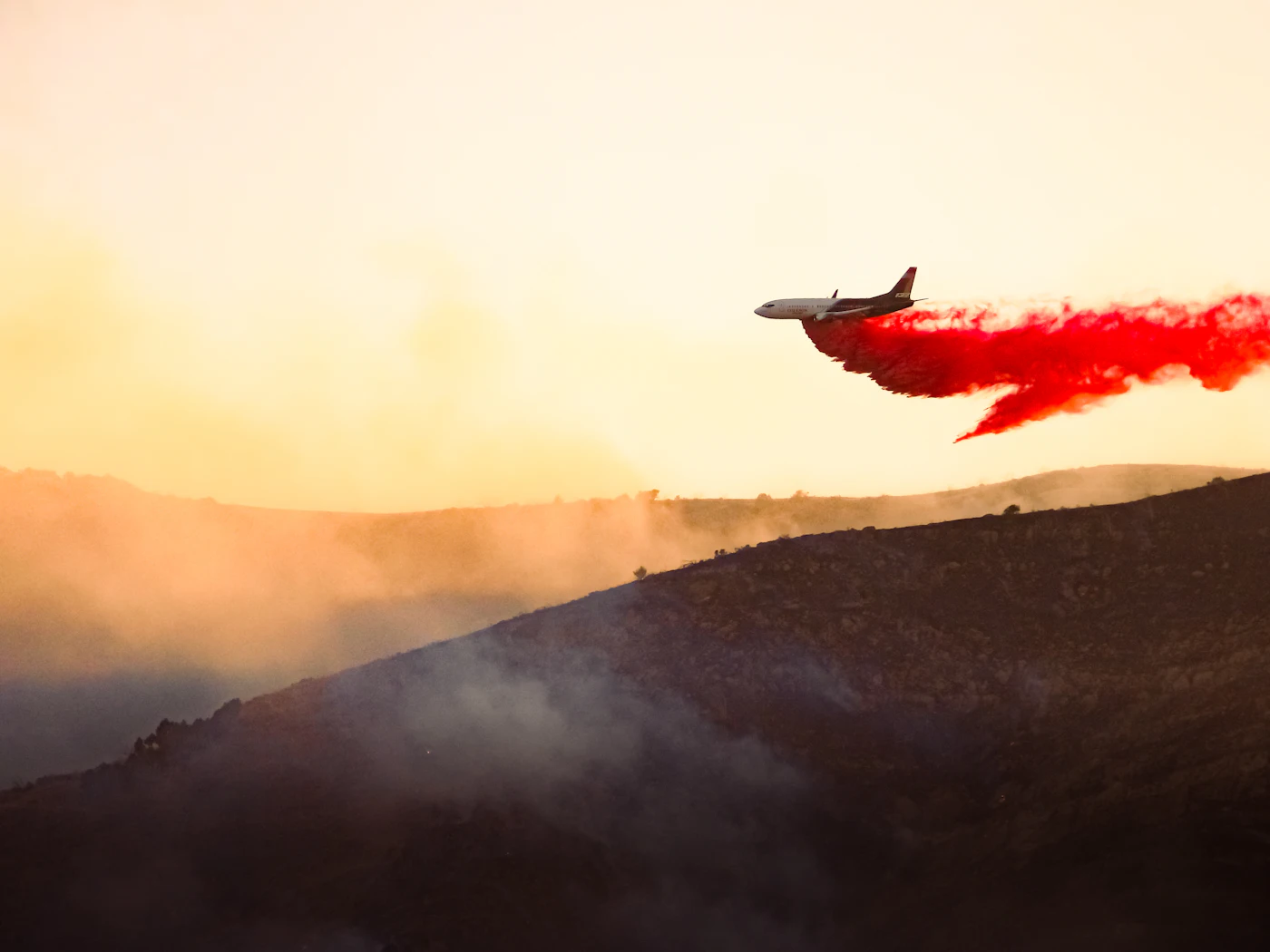 Air tanker dropping retardant over a smoke-filled valley at sunset