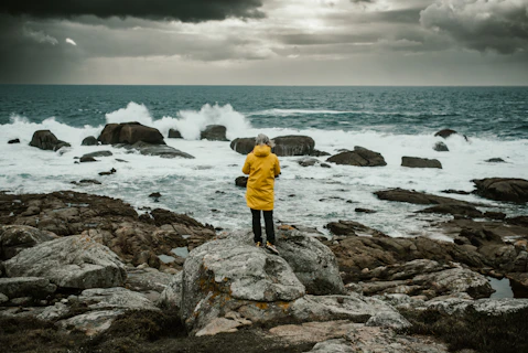 A person in a yellow jacket standing on rocks near the ocean