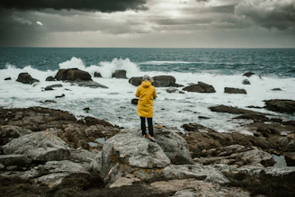A person in a yellow jacket standing on rocks near the ocean
