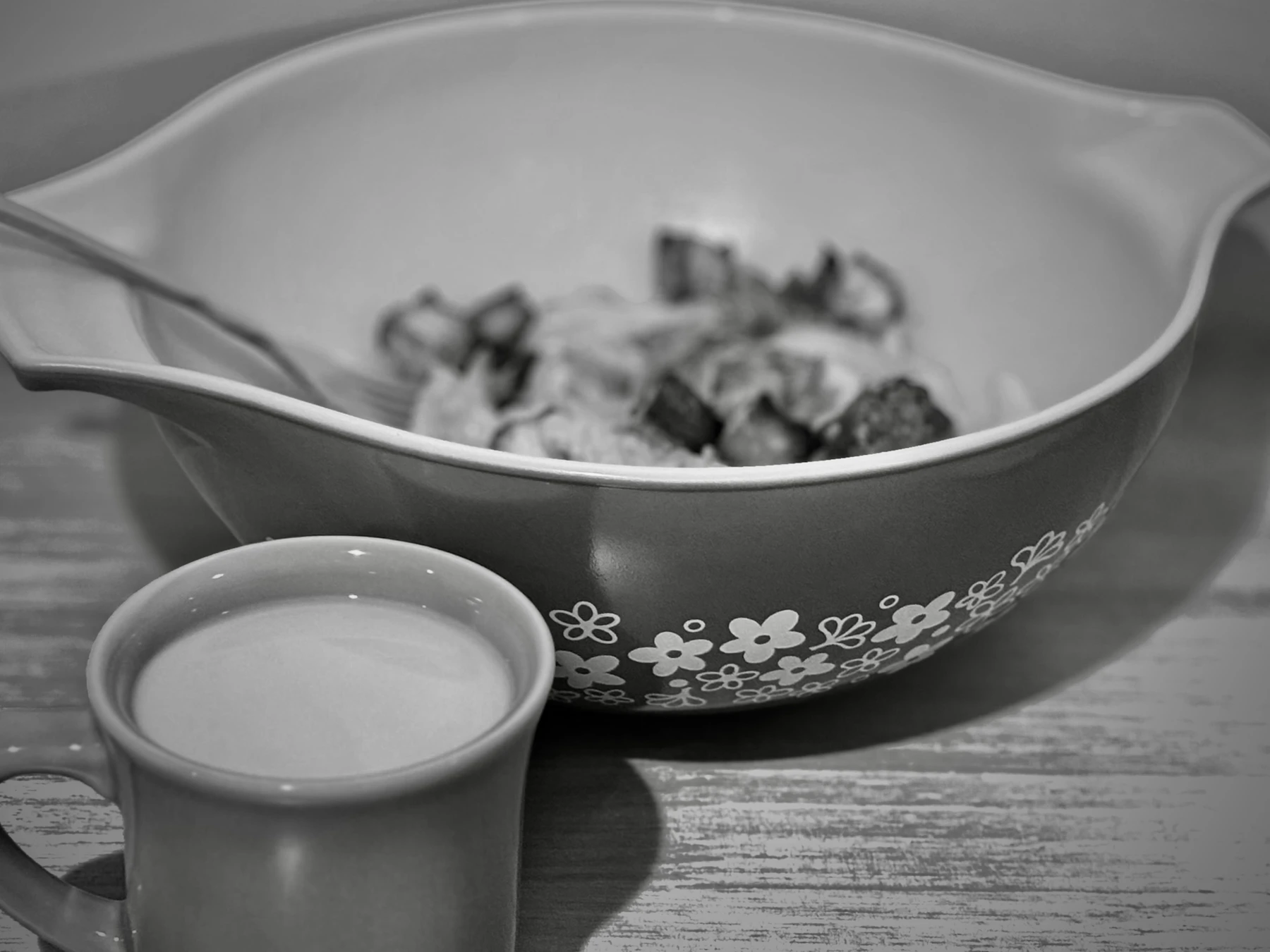 Black and white photo of a spouted glass mixing bowl with a band of printed flowers, next to a mug