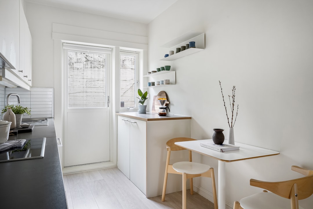 Small apartment kitchen with white cabinets, open shelving with mugs and plants, coffee station, and a two-person dining table