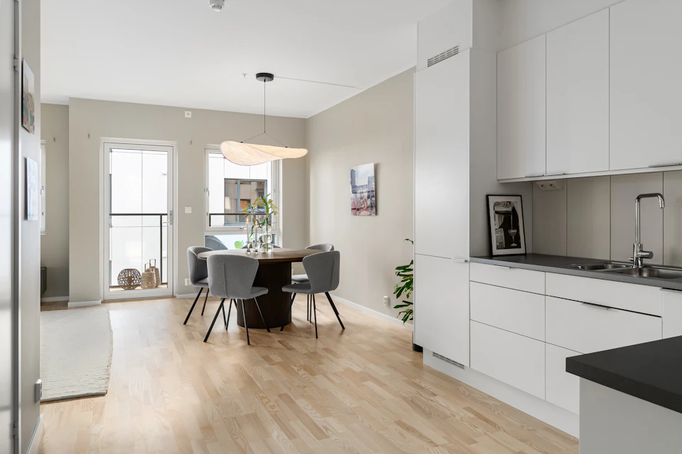 The image depicts a modern, open-concept living space with white walls and black countertops. The kitchen features white cabinets that contrast with the dark wood flooring. A large window on the left side allows natural light to flood into the room, illuminating the space. In the center of the room is a dining area with a wooden table surrounded by four chairs, two gray chairs, and a potted plant placed in front of them.

The kitchen has white appliances that blend seamlessly with the overall design. A sink can be seen on one side of the room, while a refrigerator stands tall against another wall. The space is further enhanced by a large window above the dining table, which offers an unobstructed view and allows for natural light to enter the room.