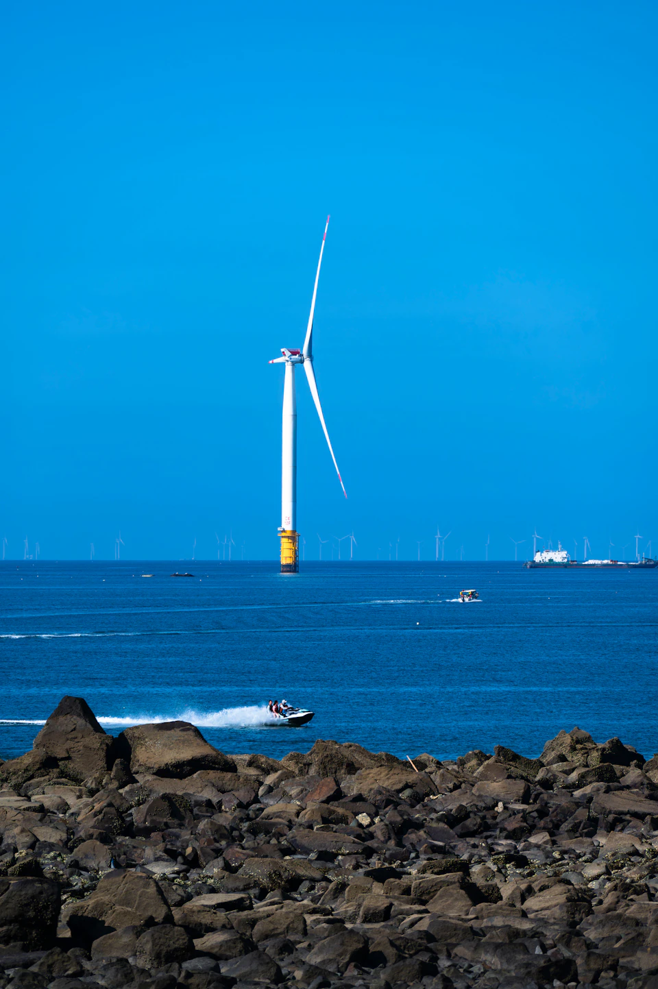 Large white wind turbine stands tall on rocky outcropping amidst smaller turbines and boats