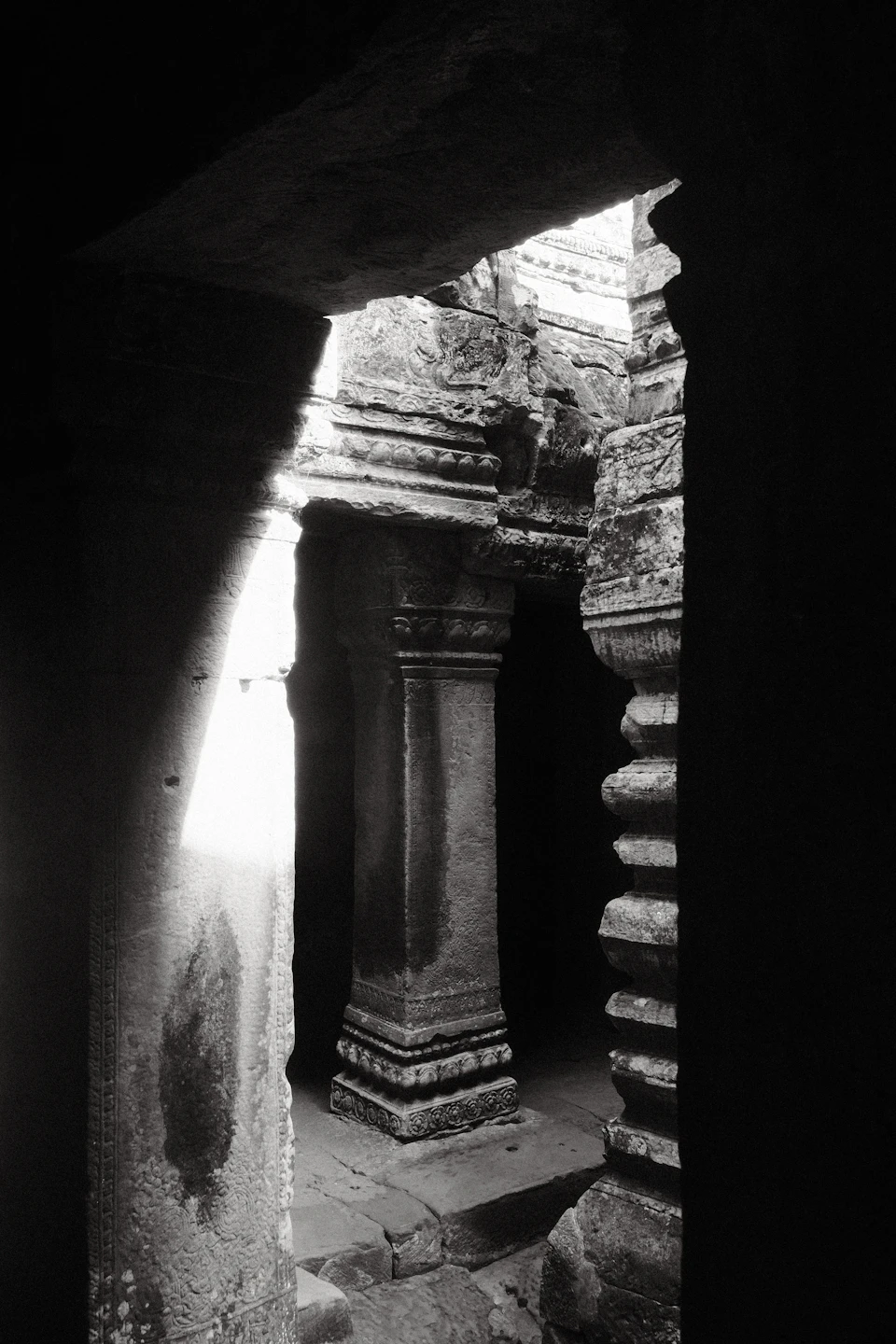 The image depicts a stone archway with two columns, one on each side, situated within an ancient temple complex. The columns are adorned with intricate carvings and patterns, showcasing the craftsmanship of the era. The perspective of the photo is taken from behind the columns, looking out through them towards the interior of the temple. The image is in black and white, adding a timeless quality to the scene.