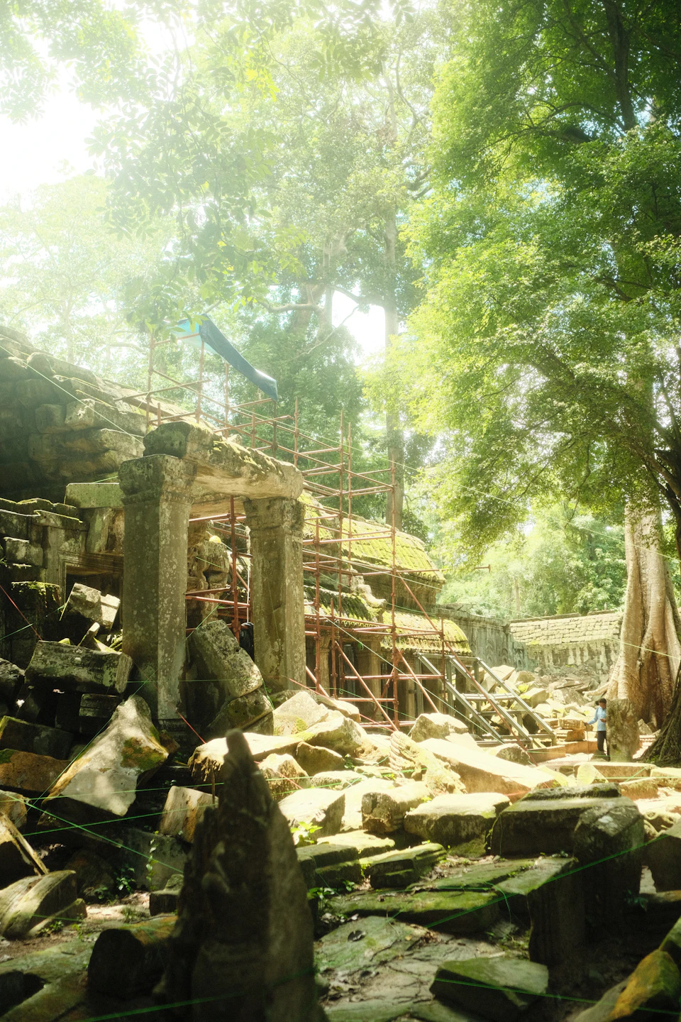 The image depicts a temple nestled within a jungle setting, with a large stone archway serving as its entrance. The archway is partially covered by scaffolding and surrounded by an array of rocks and trees. A person can be seen walking near the temple, adding a sense of scale to the scene.