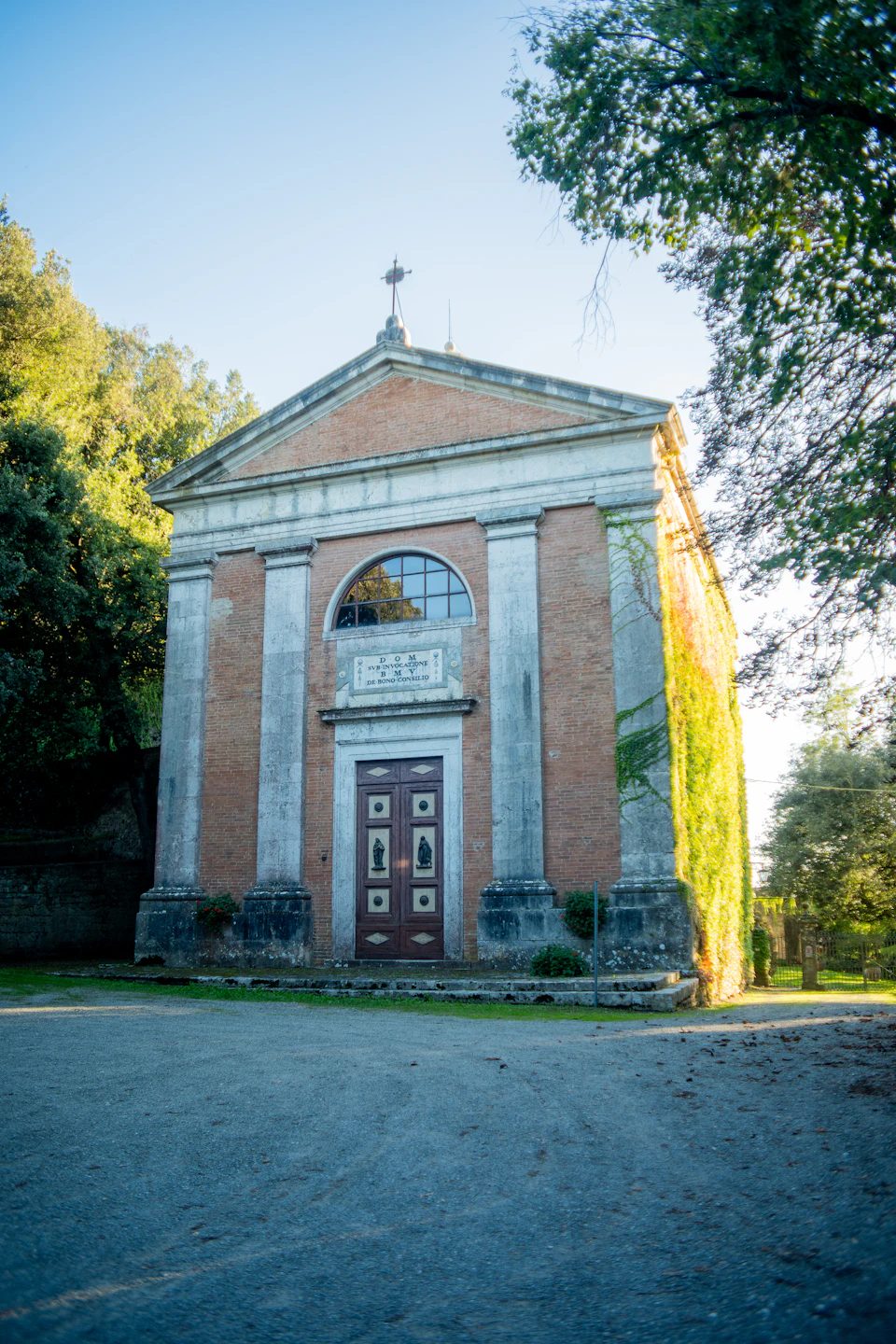 The image depicts a small, old brick building with a large arched window and a cross on top. The building is situated within a lush green landscape, surrounded by trees that provide a natural backdrop to its rustic charm. The perspective of the photo suggests it was taken from across the street or driveway, offering a clear view of the building's facade and the surrounding greenery.

The sky above is a serene blue, indicating a bright and sunny day.