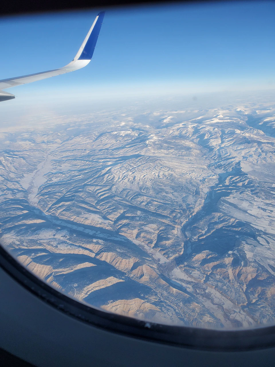 The image shows an aerial view from a plane window, capturing the breathtaking scenery below. The landscape is dominated by snow-capped mountains and valleys, with patches of green vegetation scattered throughout. A blue airplane wing can be seen on the left side of the frame, adding to the sense of scale and grandeur of the scene.