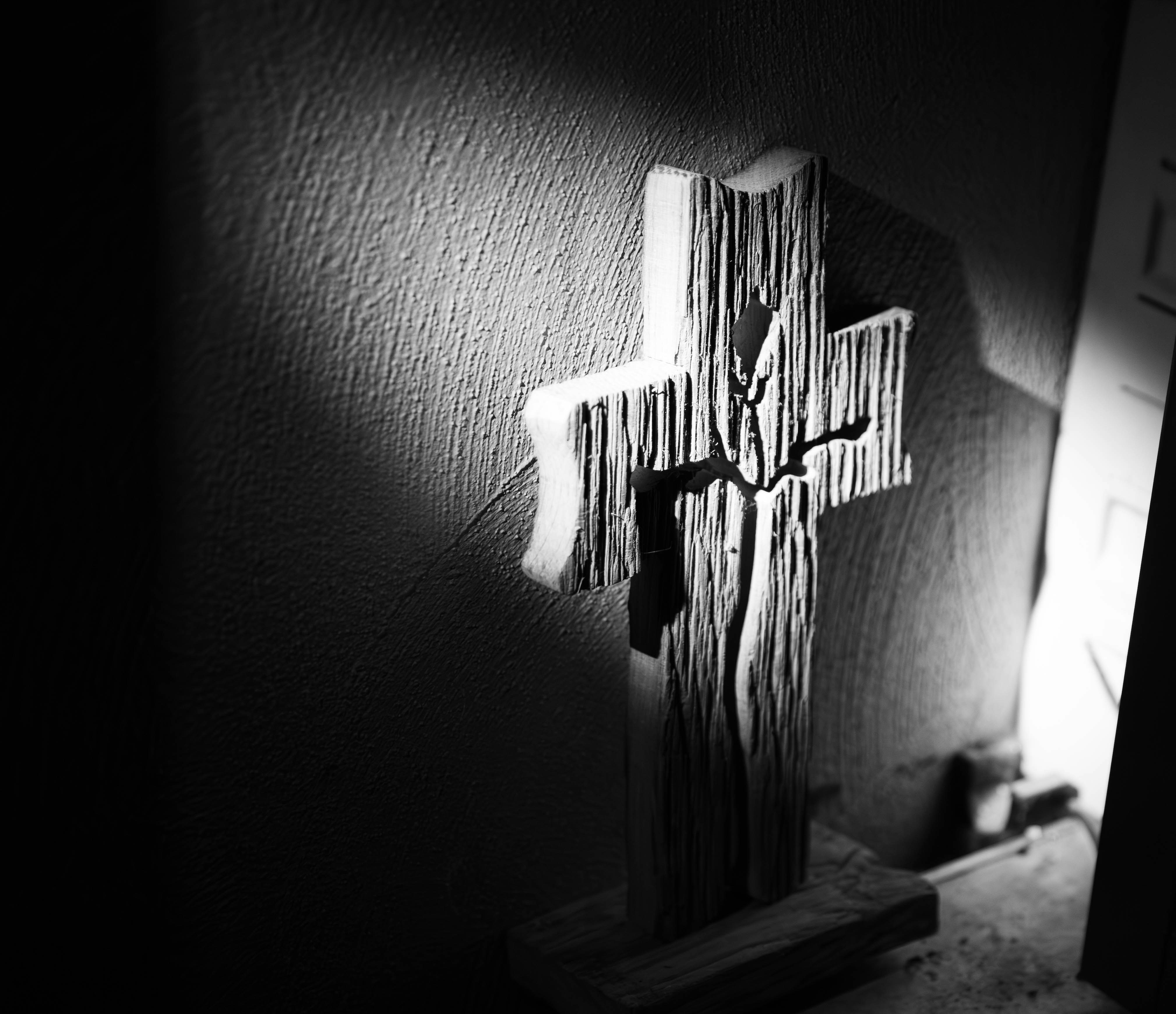 Rustic wooden cross mounted on a warm white plaster wall, golden hour sunlight casting soft shadows, natural wood grain texture