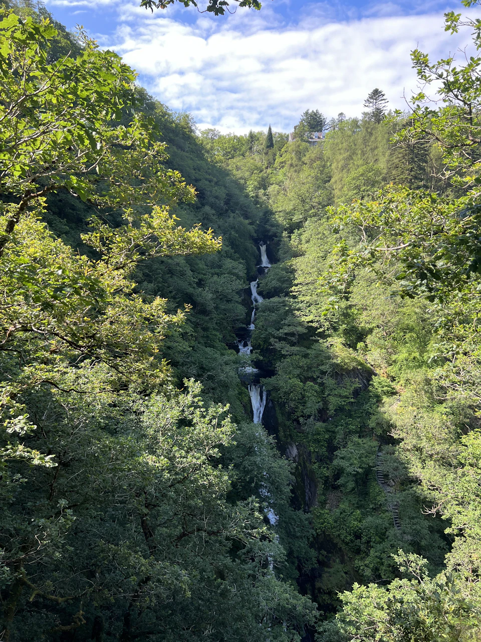 The image depicts a serene forest scene with a tall, narrow waterfall cascading down into a valley below. The waterfall is surrounded by lush green trees and bushes, creating a sense of tranquility and natural beauty. The perspective of the photo is from above, providing an expansive view of the waterfall and its surrounding landscape.