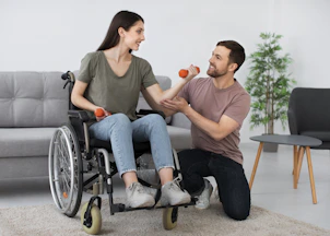 A woman in a wheelchair lifting orange dumbbells with a man kneeling beside her