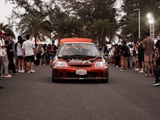 A red car driving down a street next to a crowd of people