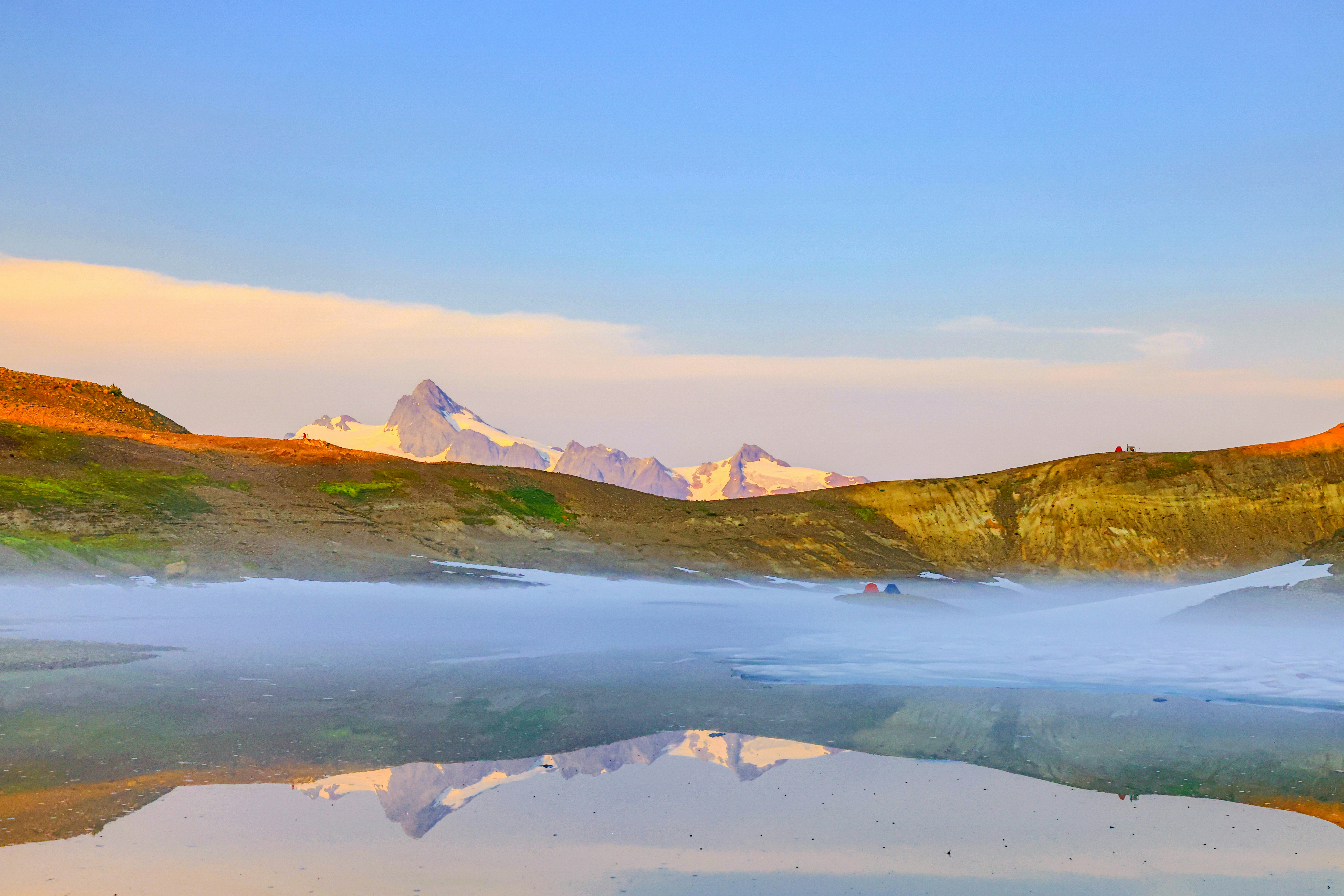 A tranquil lake reflecting the surrounding mountains and the sky, capturing the stillness of a serene moment in the highlands. The soft, pastel colors of the sky suggest the peacefulness of dawn or dusk.
