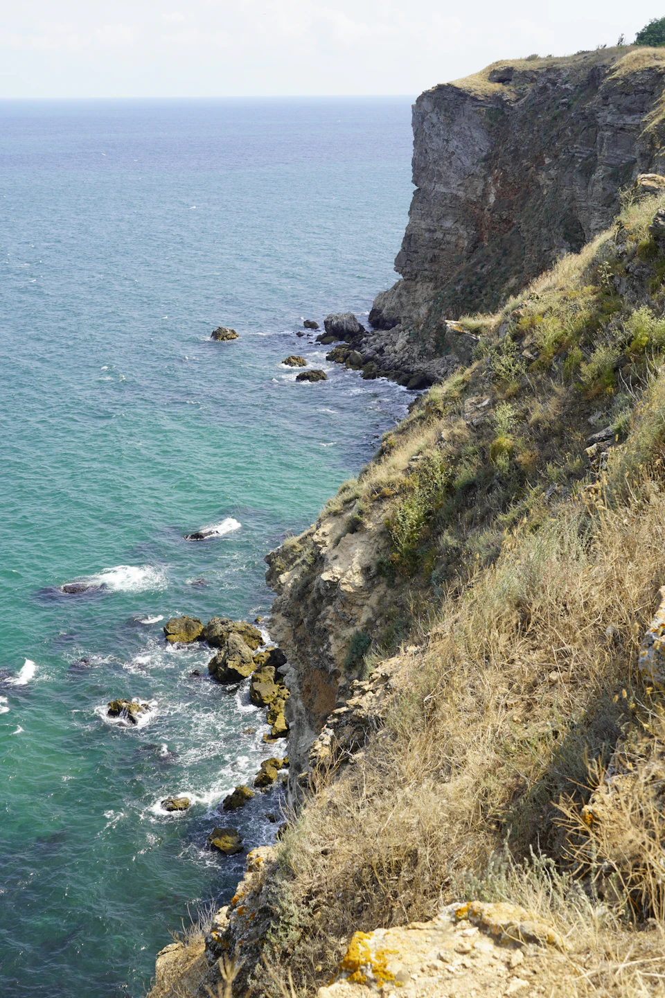 The image depicts a cliff overlooking the ocean, with a rocky coastline and a grassy area on one side. The cliff is covered in green vegetation, contrasting with the brown hues of the rocks. The deep blue-green water below reflects the sky above, creating a serene atmosphere. The perspective of the photo suggests it was taken from the top of the cliff, offering a panoramic view of the landscape and the vast expanse of the ocean beyond.