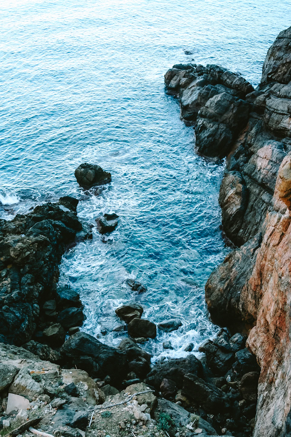 Rocky cliff overlooking deep blue ocean with large boulders and contrasting vibrant water below