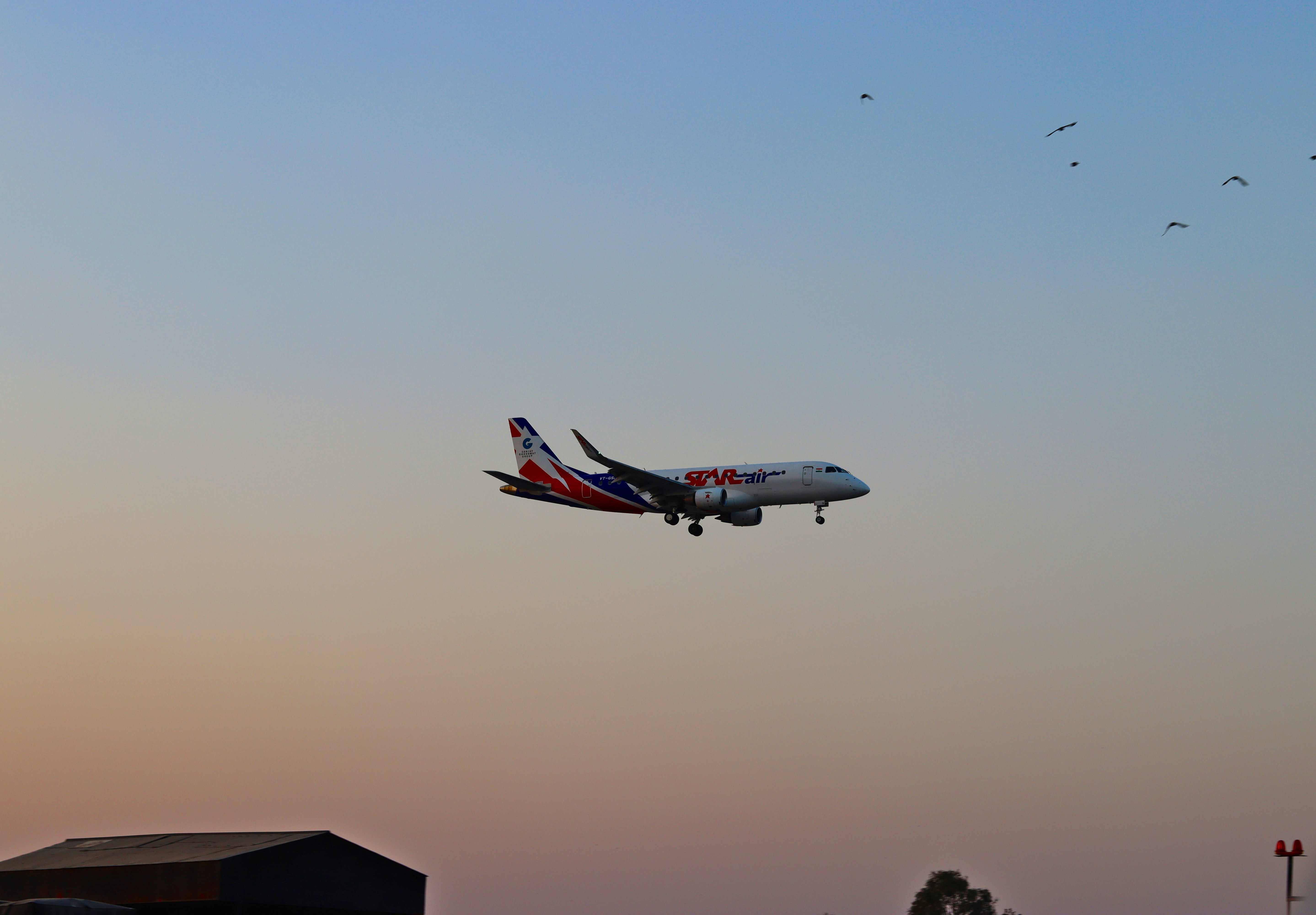 An airplane is flying over a field at sunset, A Star Air Embraer E175 near HAL Airport, Bengaluru