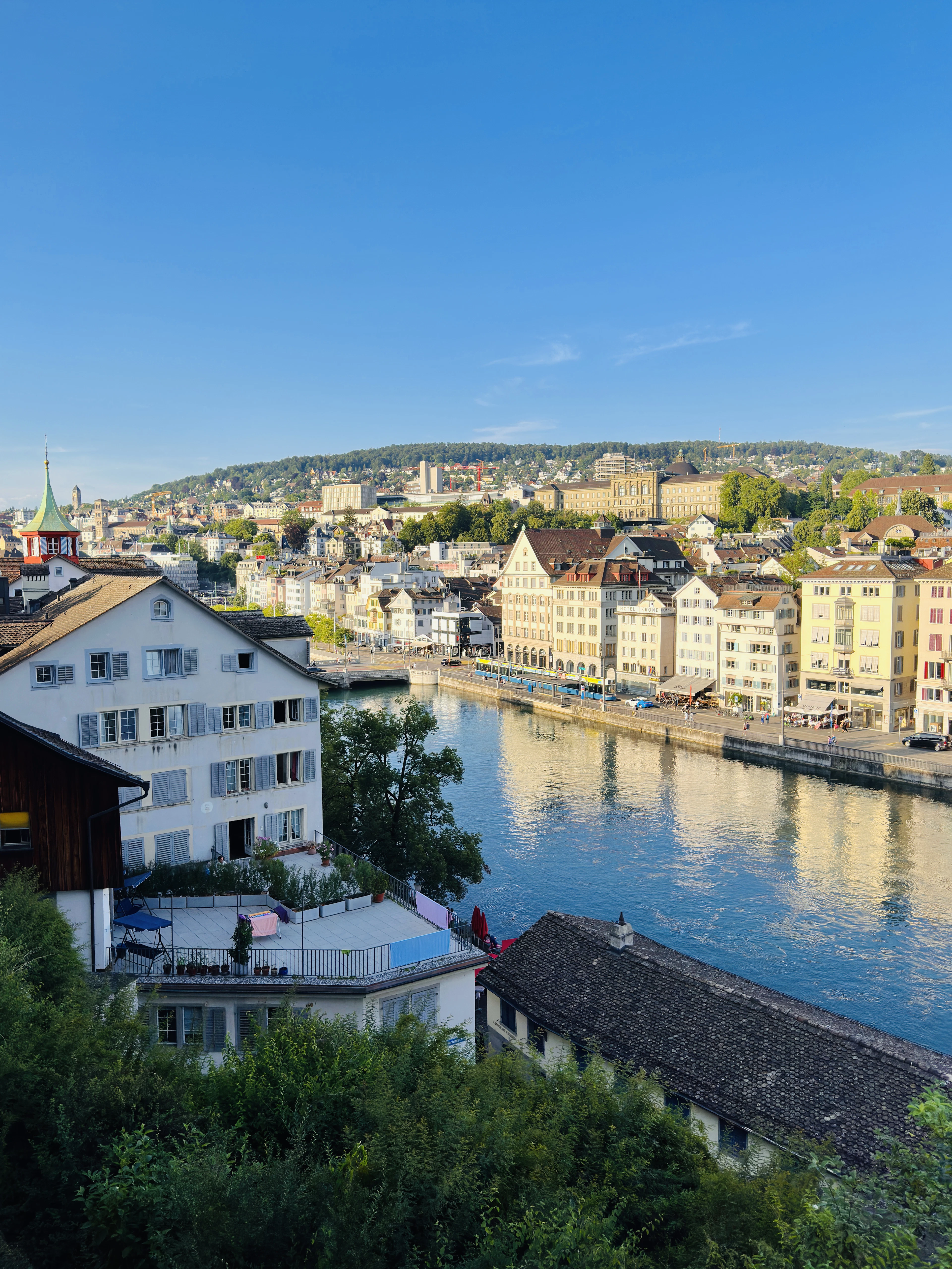 A hillside view over the Limmat river shows old-town rooftops, a church spire, and green hills beyond.