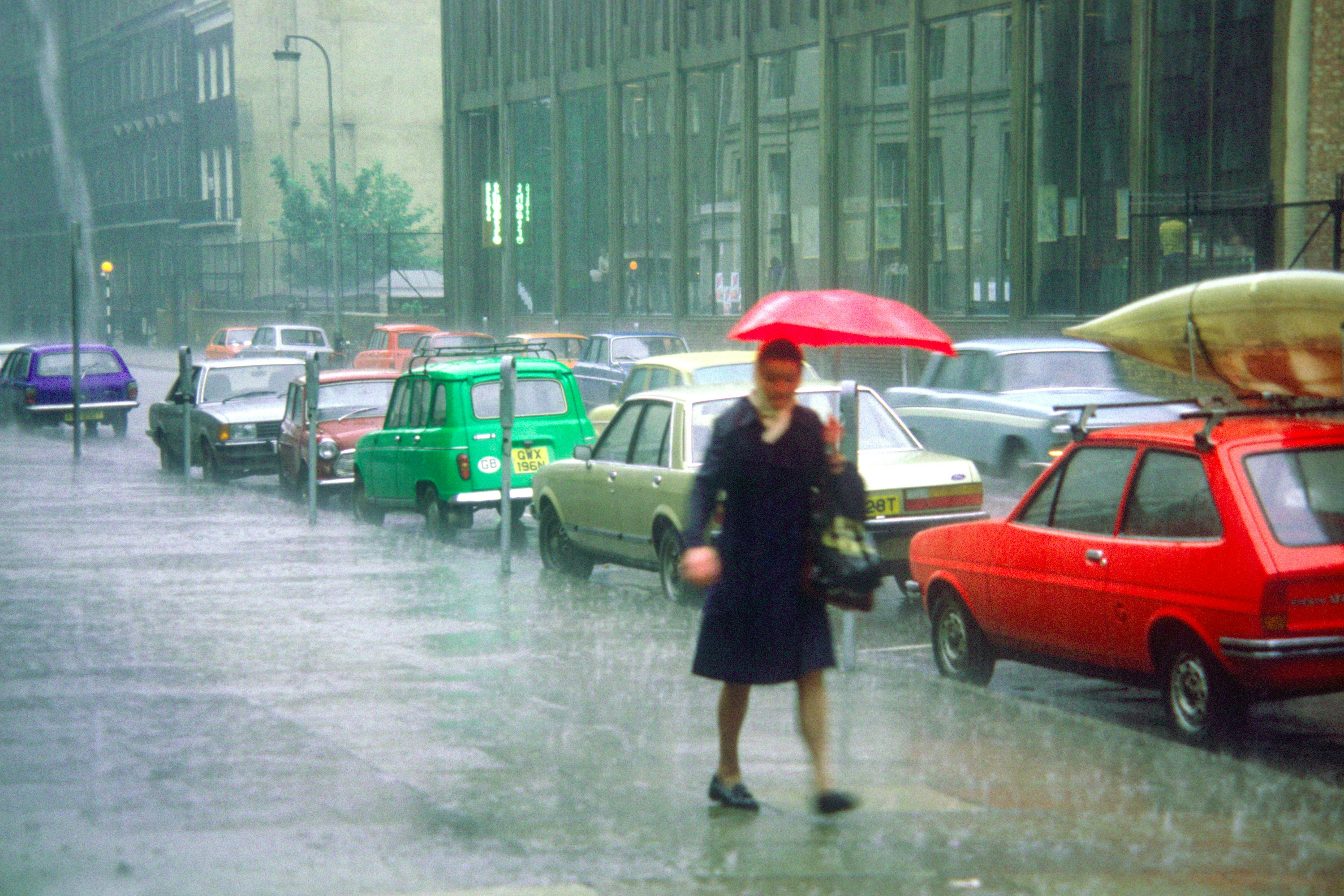 woman with red umbrella in downpour. sidewalk with cars on street
