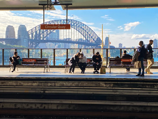 Commuters wait on the Circular Quay railway platform with the Sydney Harbour Bridge visible beyond the station sign.