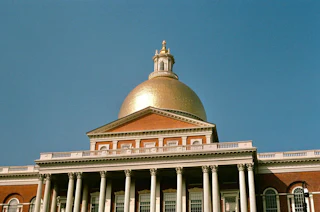 Gold dome and white Corinthian columns of the Massachusetts State House against a clear blue sky