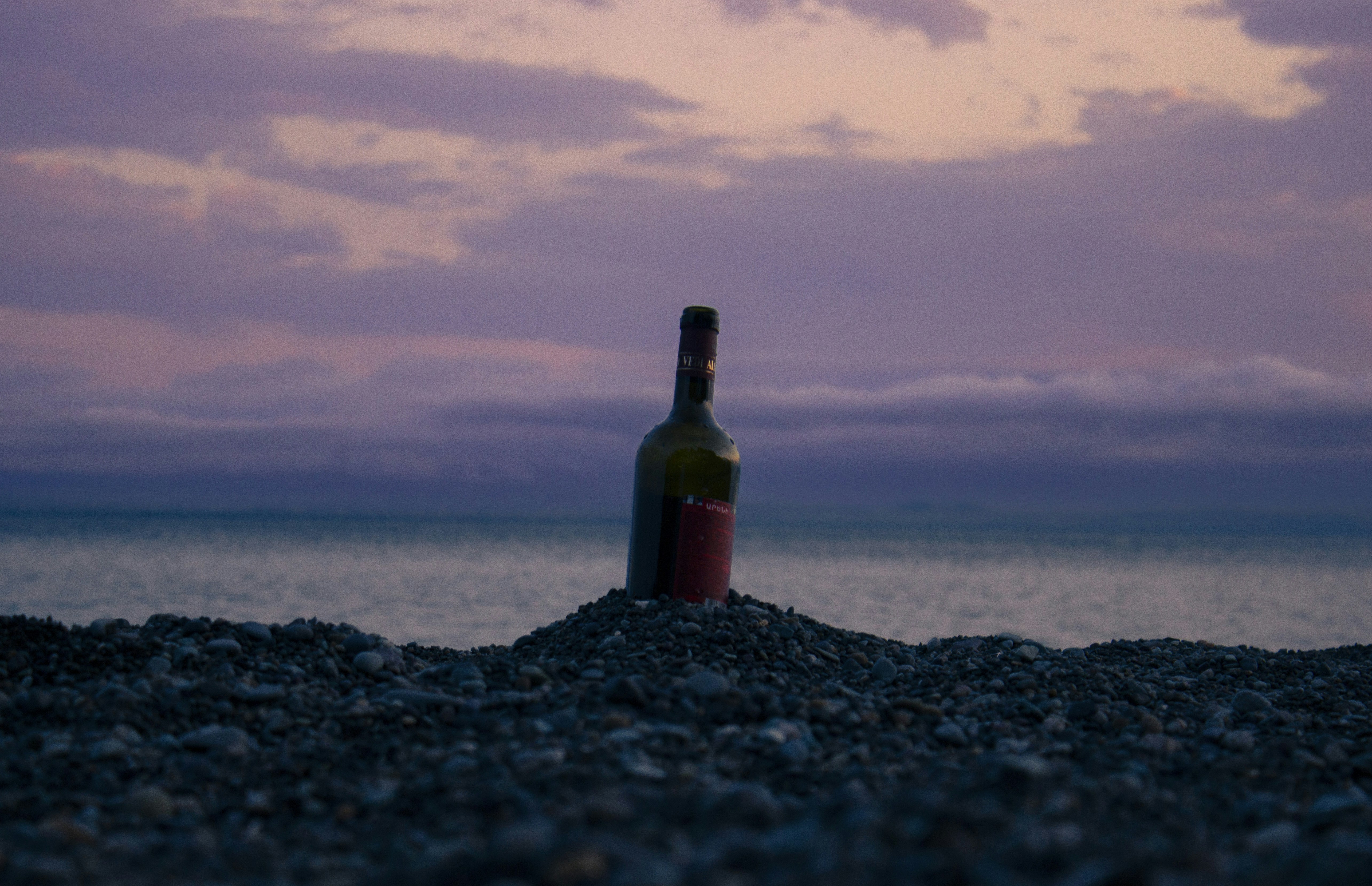 A bottle of wine sitting on top of a rocky beach, Scenic view of a bottle half buried on the beach on the shore at a beautiful pink cloudy sunset