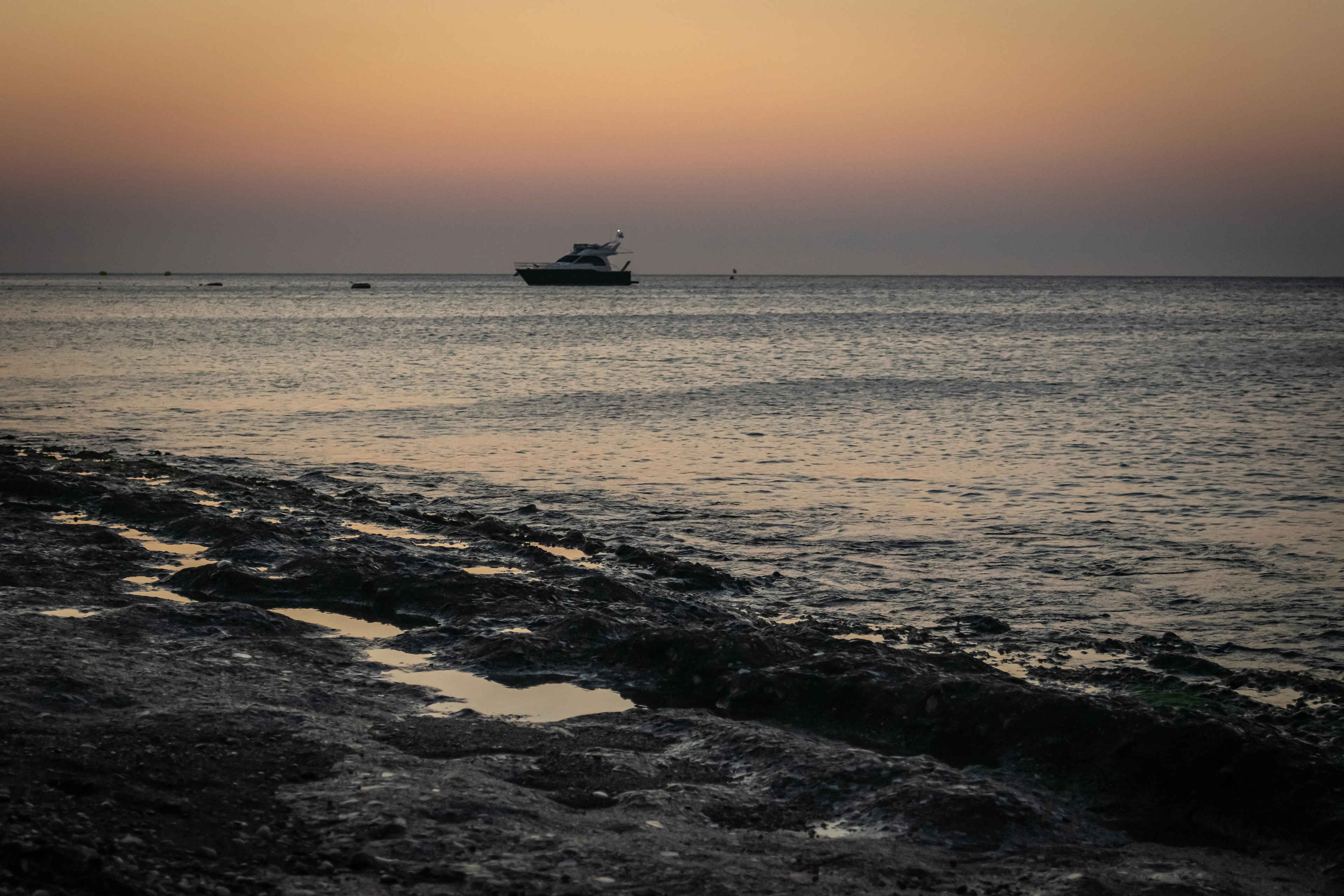 A small boat anchored on a calm sea glowing orange at sunset, seen from a rocky Rhodes shoreline.