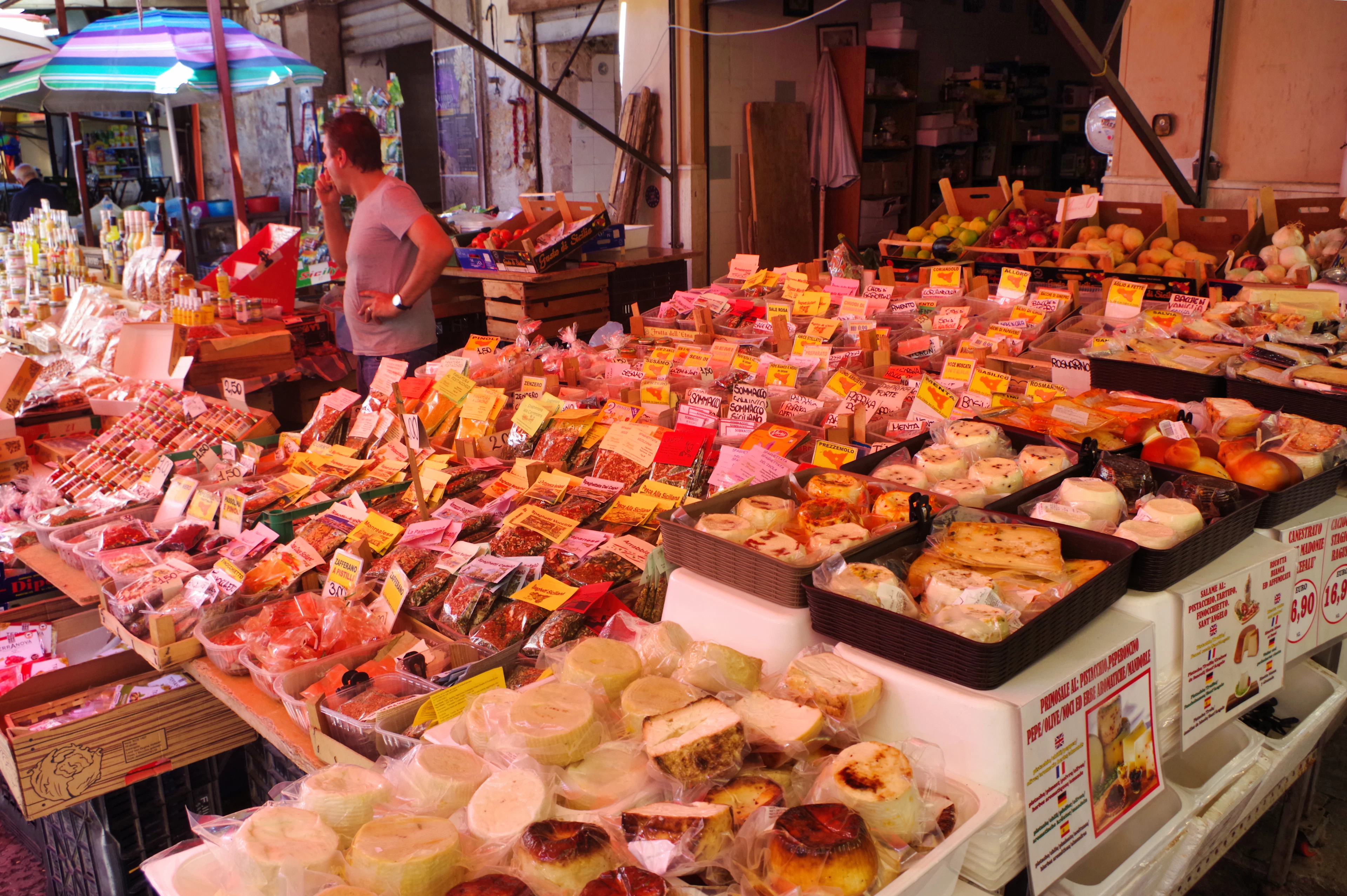 A street market stall piled high with cheeses, cured meats and prepared foods under colorful awnings