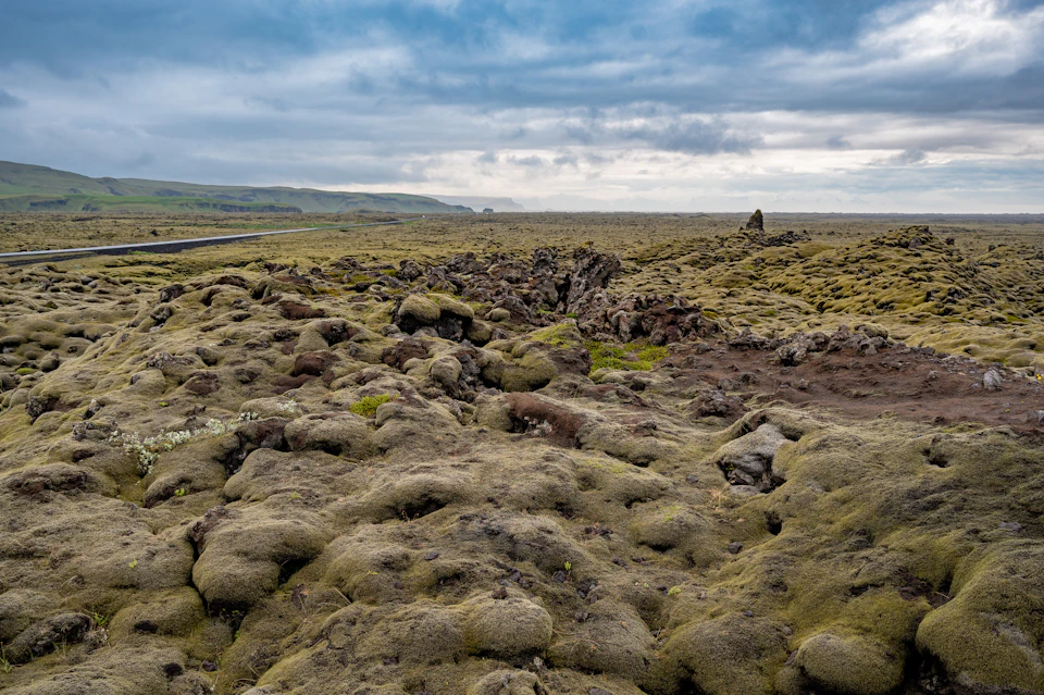 The image depicts a vast, barren landscape with a rocky terrain and patches of greenery scattered throughout. The sky above is filled with clouds, casting a soft light over the scene. In the distance, there are hills that add depth to the landscape. A road can be seen in the background, suggesting human presence or activity in this remote area.
