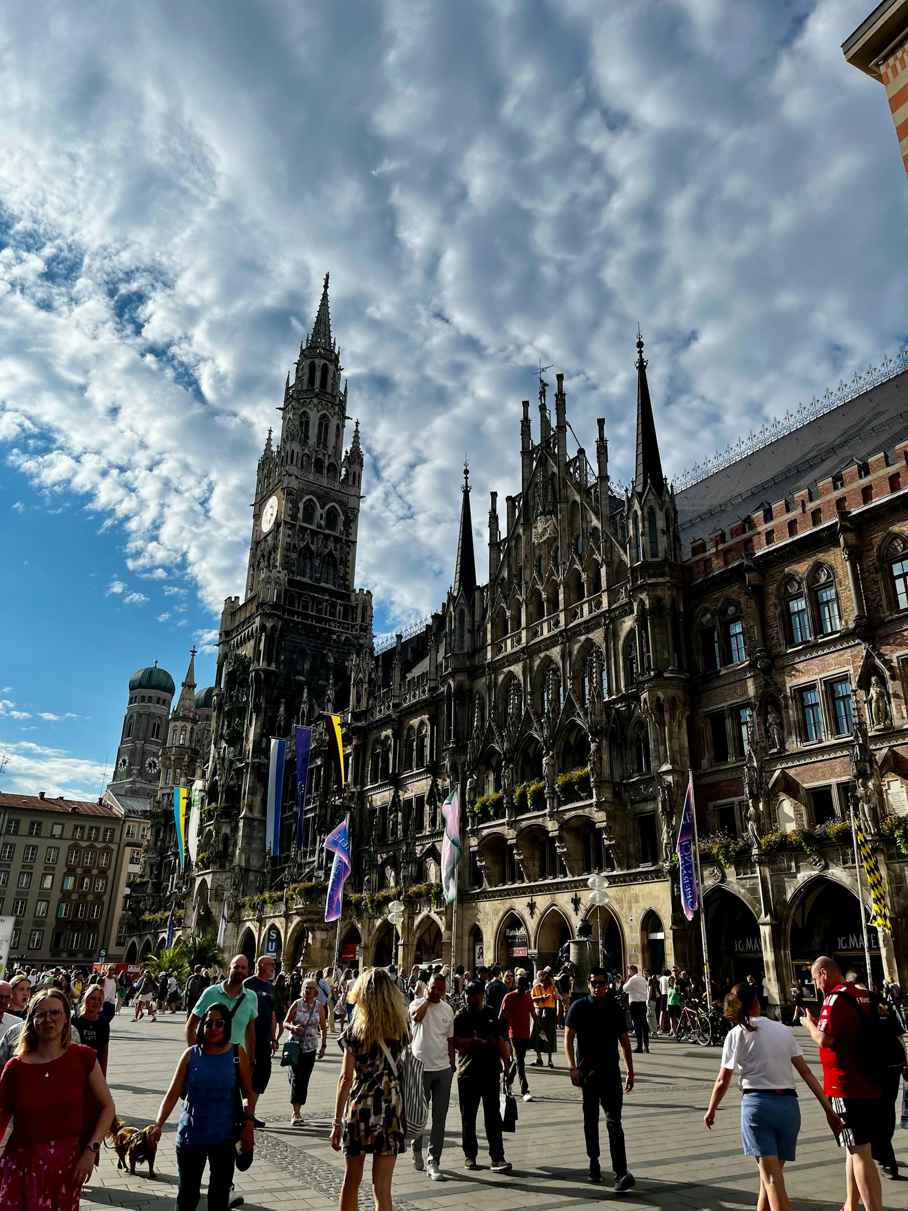 People in Munich’s Marienplatz beneath an ornate stone facade and cloudy sky.