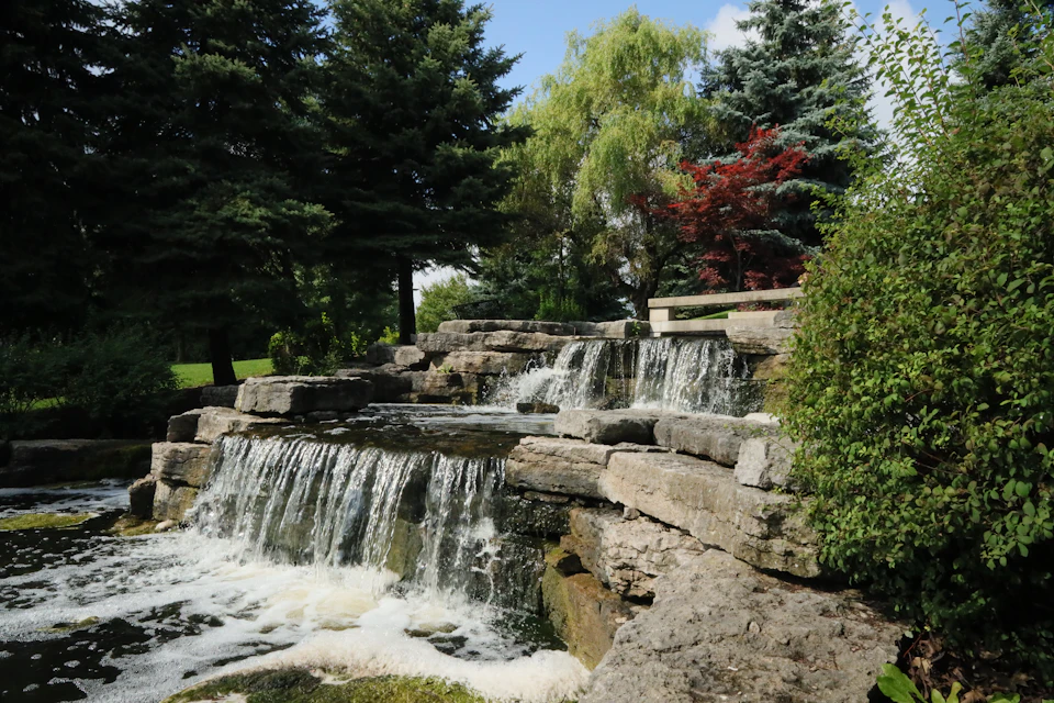 The image depicts a serene park scene with a cascading waterfall composed of large rocks and boulders, surrounded by lush greenery. The waterfall is situated on the right side of the frame, creating a sense of depth and perspective in the composition. In the background, there are trees and bushes that add to the natural beauty of the setting.