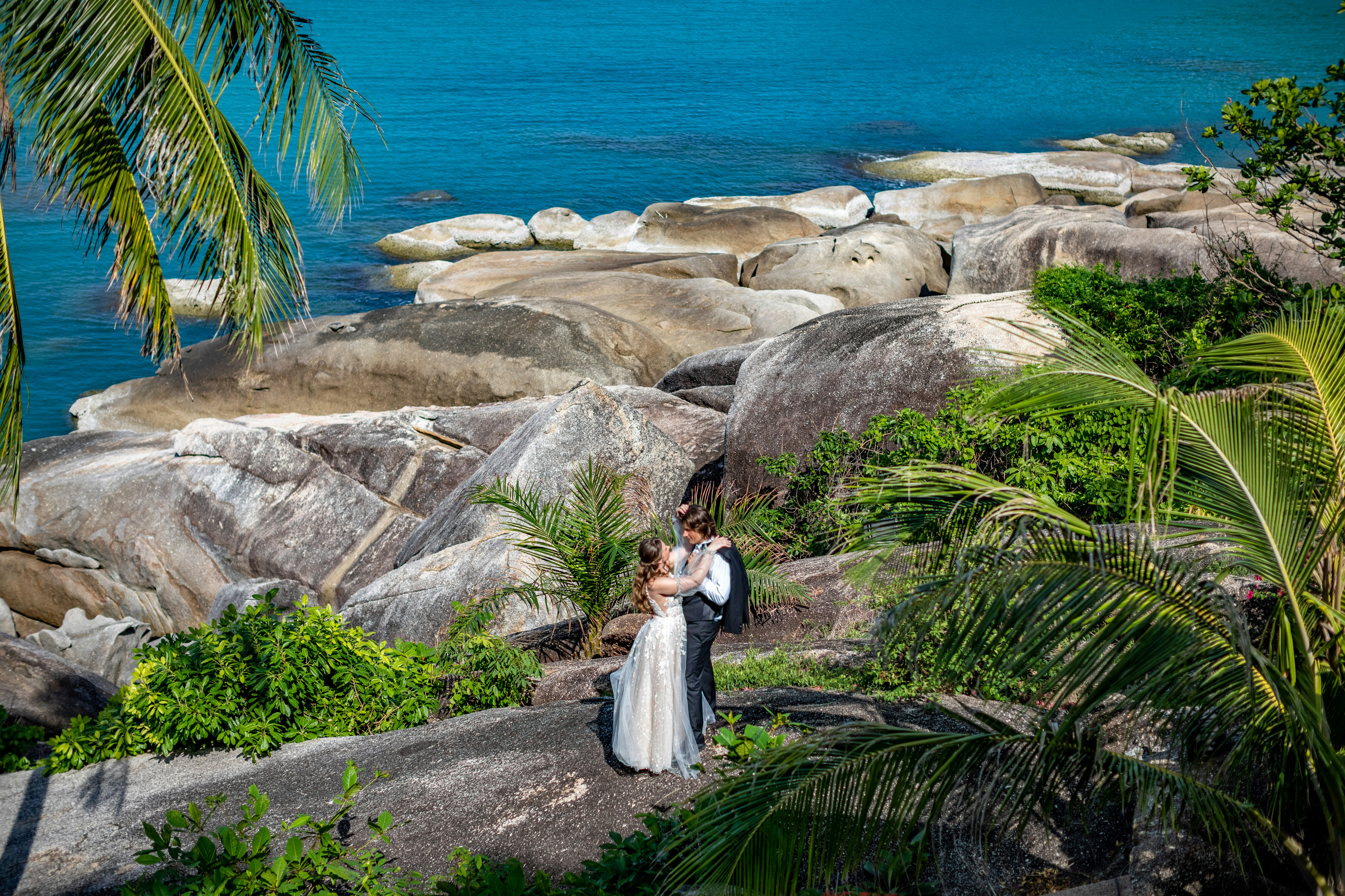 A bride and groom standing on a rock by the ocean, 