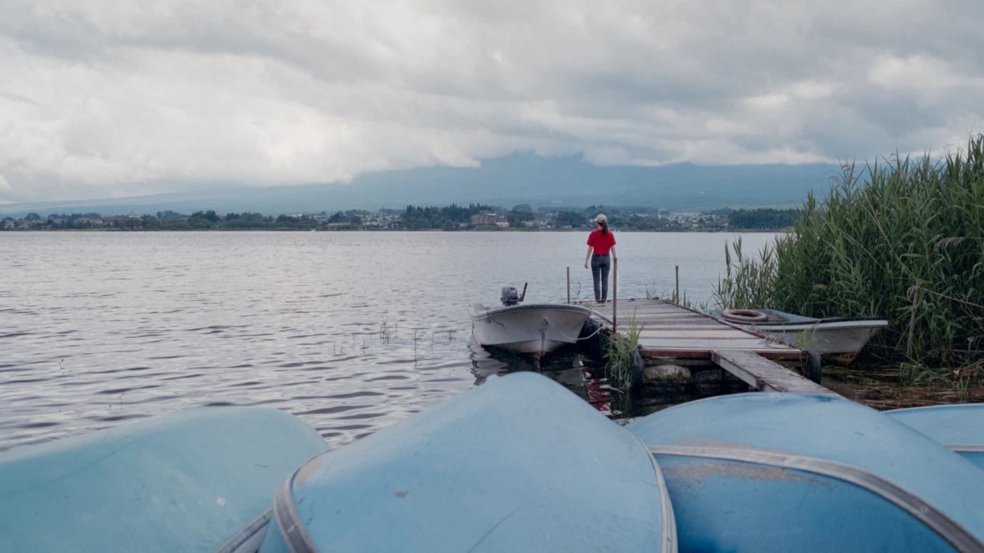 A man stands on a wooden dock beside a boat on a calm lake under a grey sky