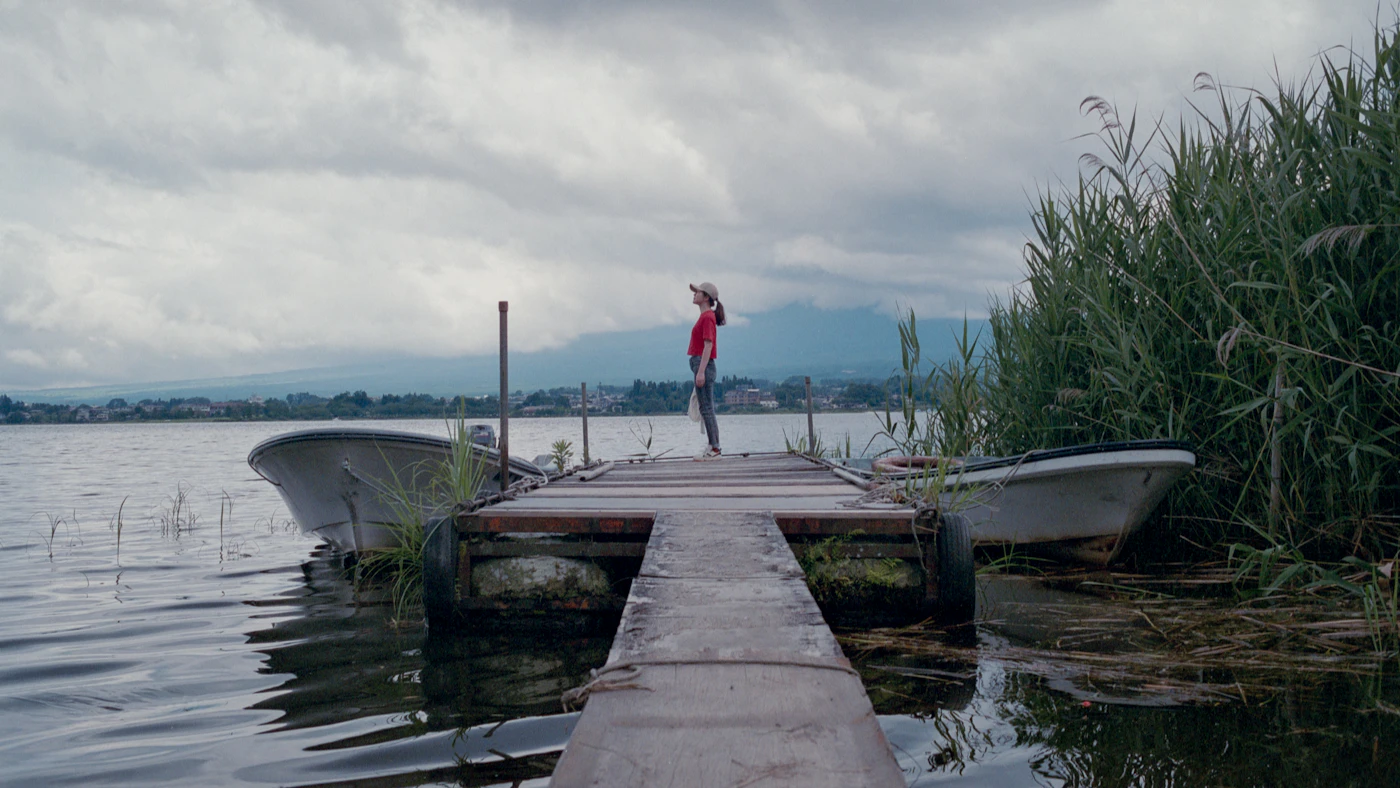 A person stands on a small wooden dock between two boats on a calm lake with reeds on the right