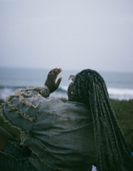 A woman with dreadlocks sitting on the grass looking at the ocean