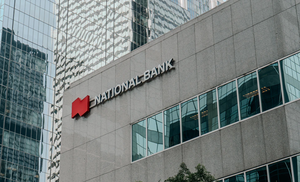 The image shows a large, modern building with a gray facade and multiple windows reflecting the surrounding cityscape. The National Bank logo is prominently displayed on the side of the building, indicating its affiliation with the financial services institution. In front of the building, there are trees providing some greenery to the urban setting.