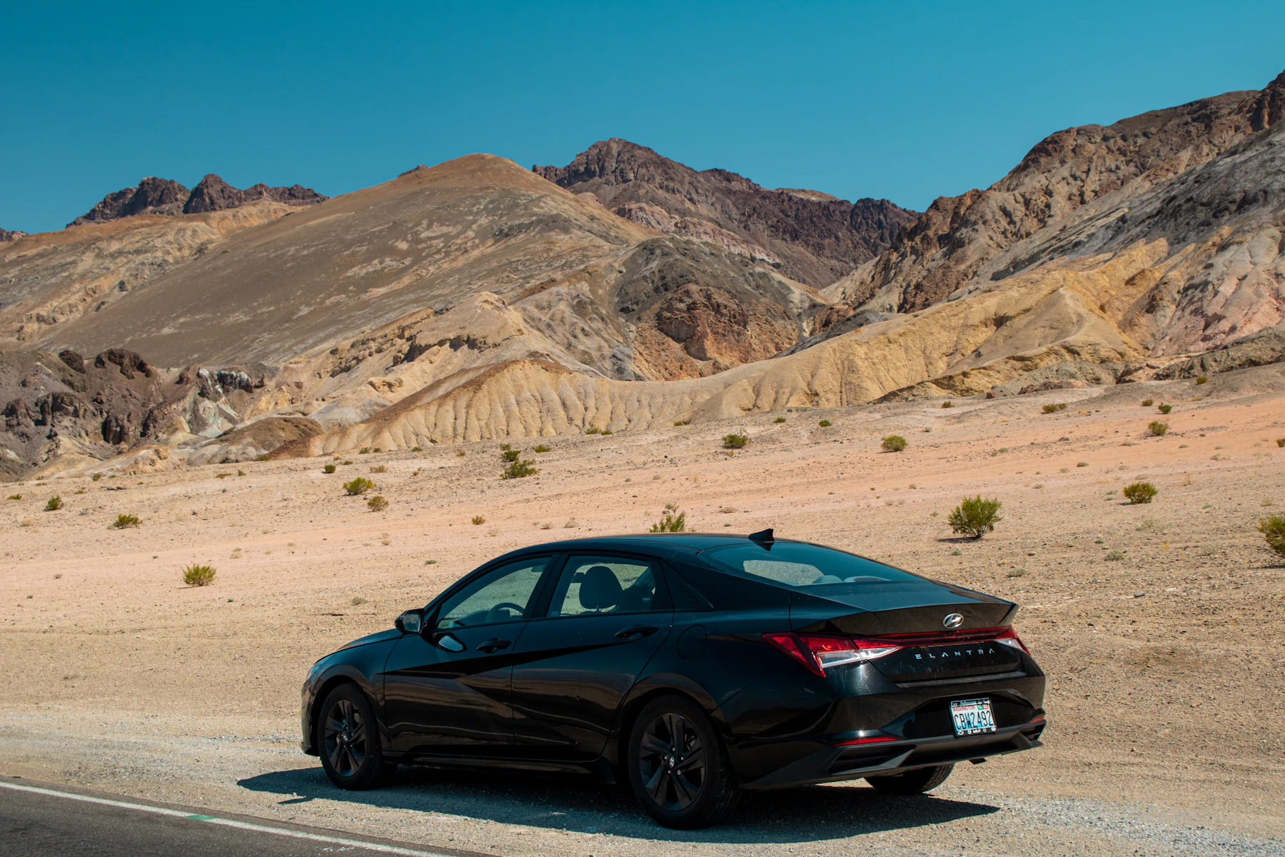 A black sedan pulled onto the gravel shoulder of a desert road below striped tan and rust hills