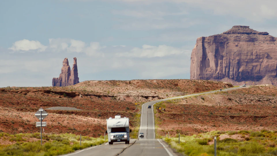 White RV travels on winding desert road past red rock formations in clear blue sky
