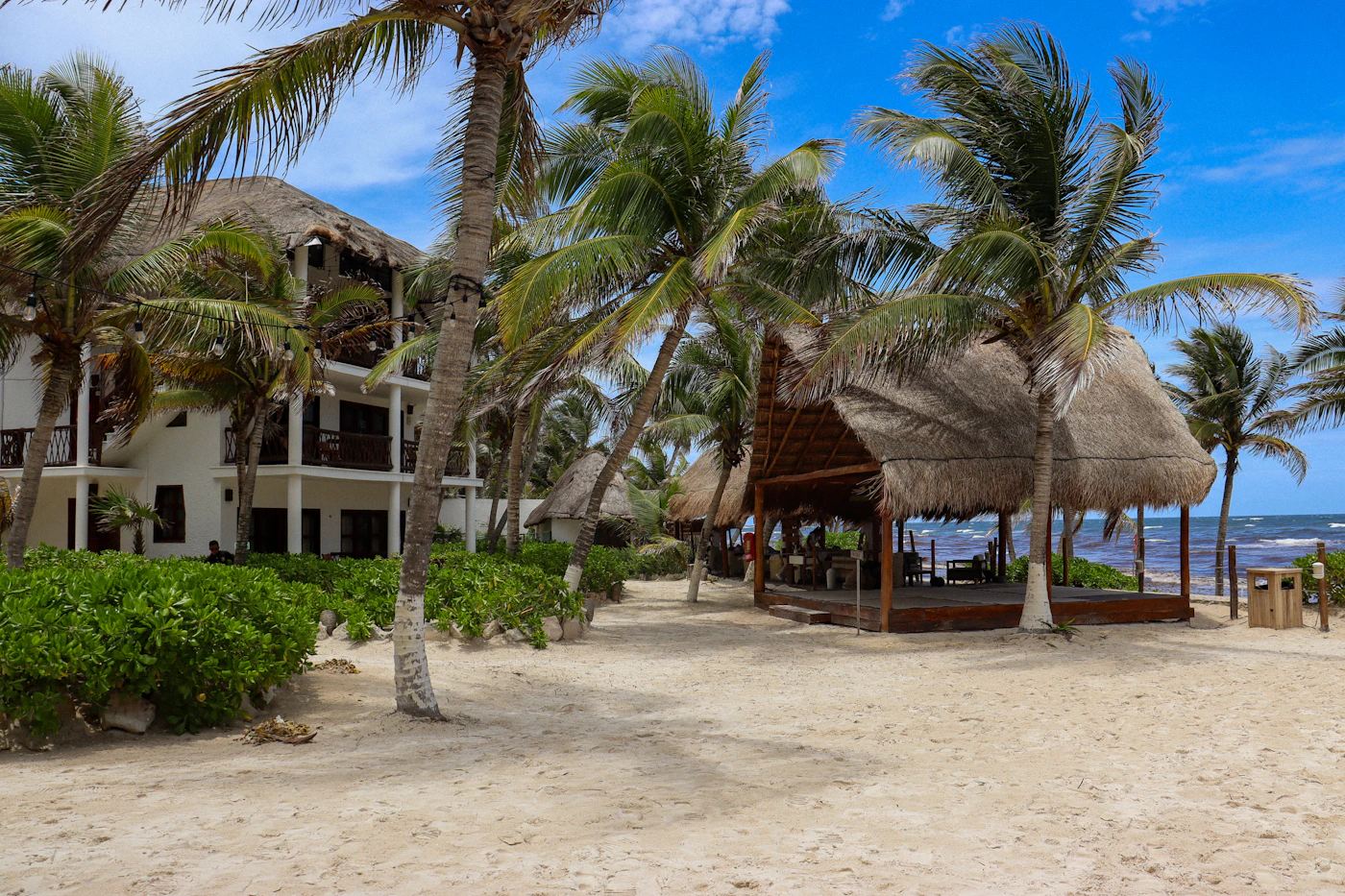 A Tulum beachfront resort: a large thatched-roof palapa open-air structure on white sand with a white villa building behind it, coconut palms throughout, turquoise Caribbean visible beyond