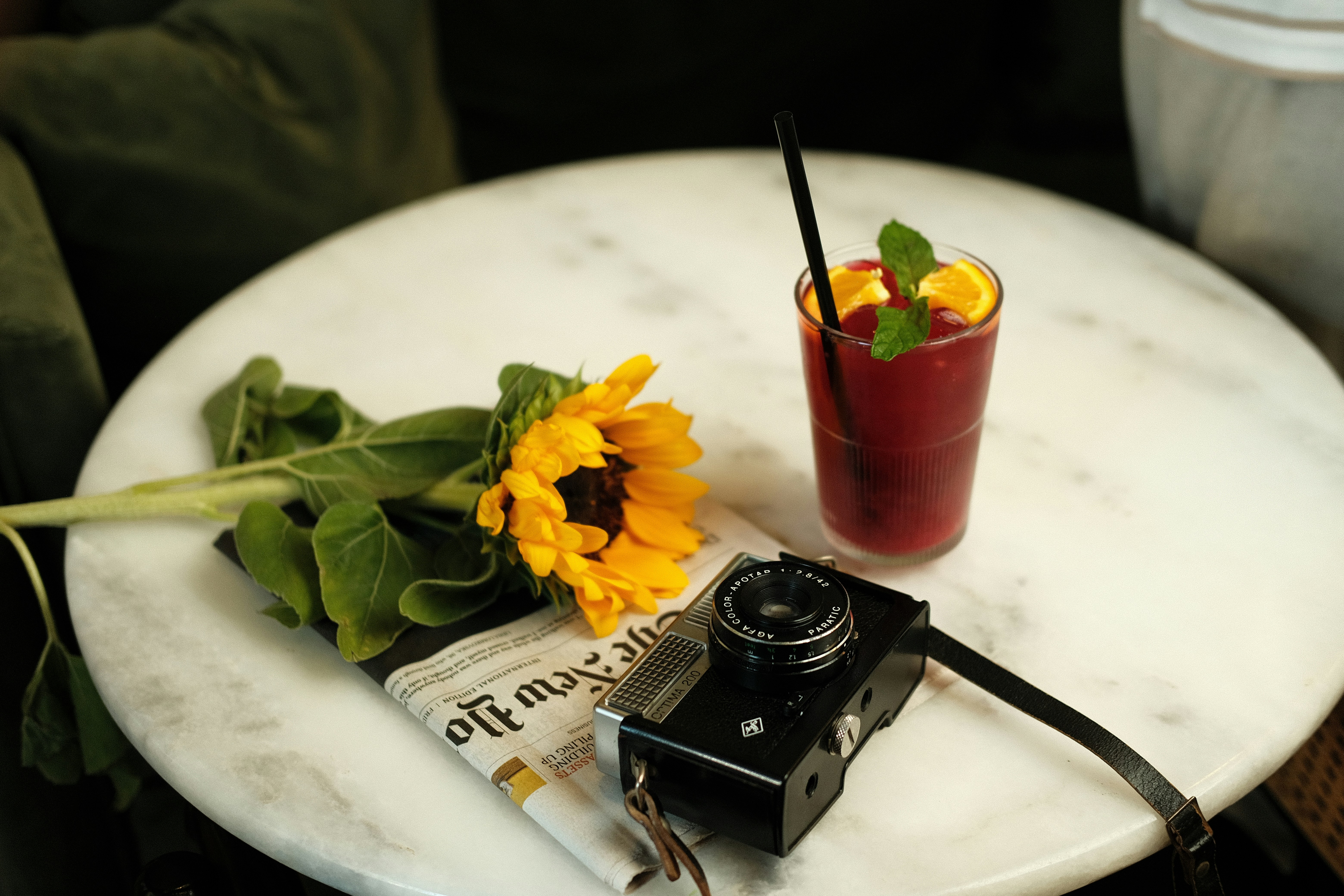 A vintage rangefinder rests on a marble table beside a bright red cocktail and a sunflower, with a folded newspaper peeking beneath. A still-life setup framed for a moody, tactile moment.