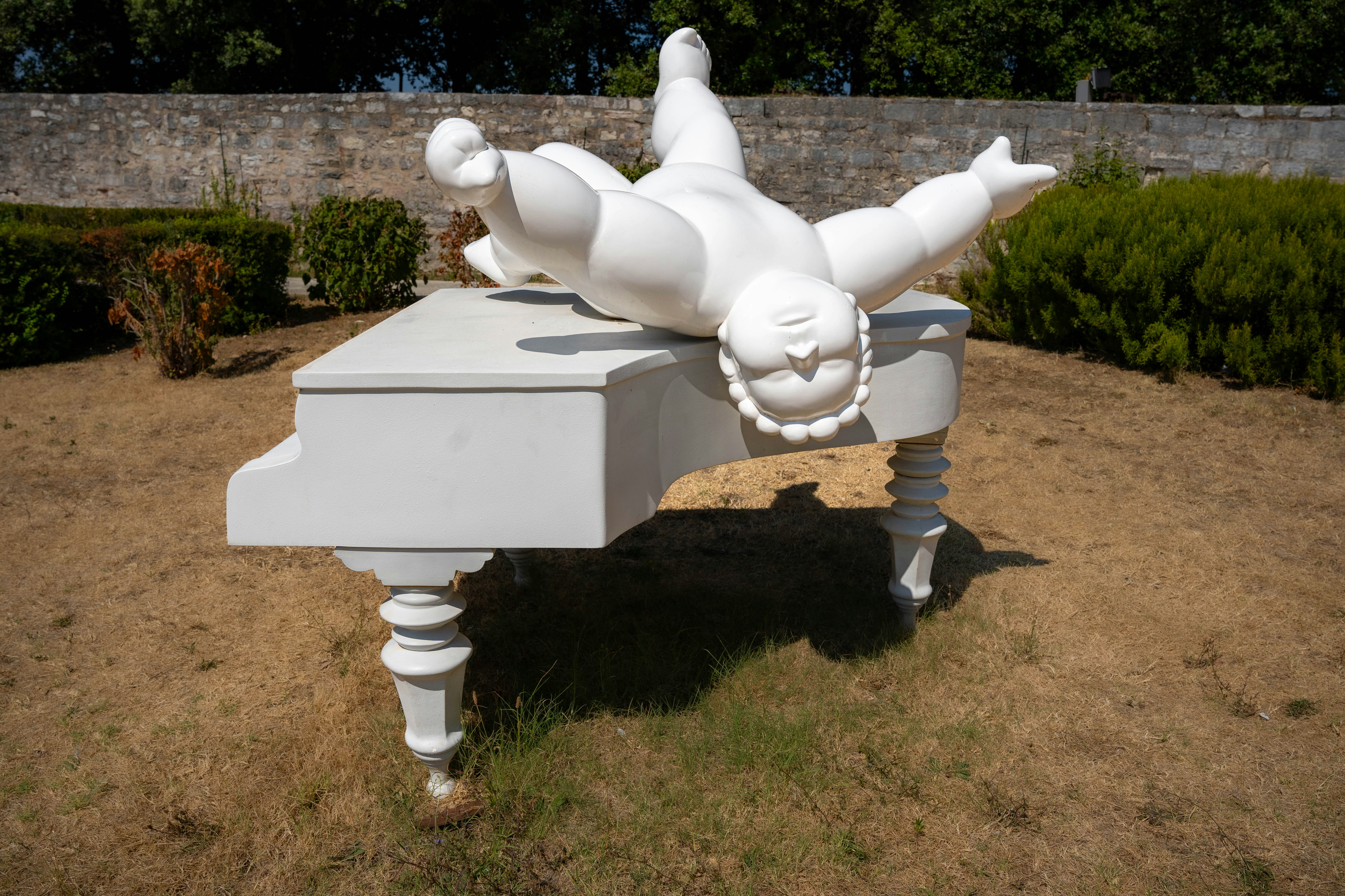 A sculpture of a person falling off a piano, The image showcases a unique and whimsical sculpture displayed outdoors on a patch of dry grass. The sculpture consists of a white, oversized figure resembling a cherubic child lying face-down on a large white grand piano. The figure’s arms and legs are spread out in a carefree or exaggerated pose, giving it a humorous and surreal appearance. The sculpture is situated in a garden or park area, with some greenery and a stone wall in the background.