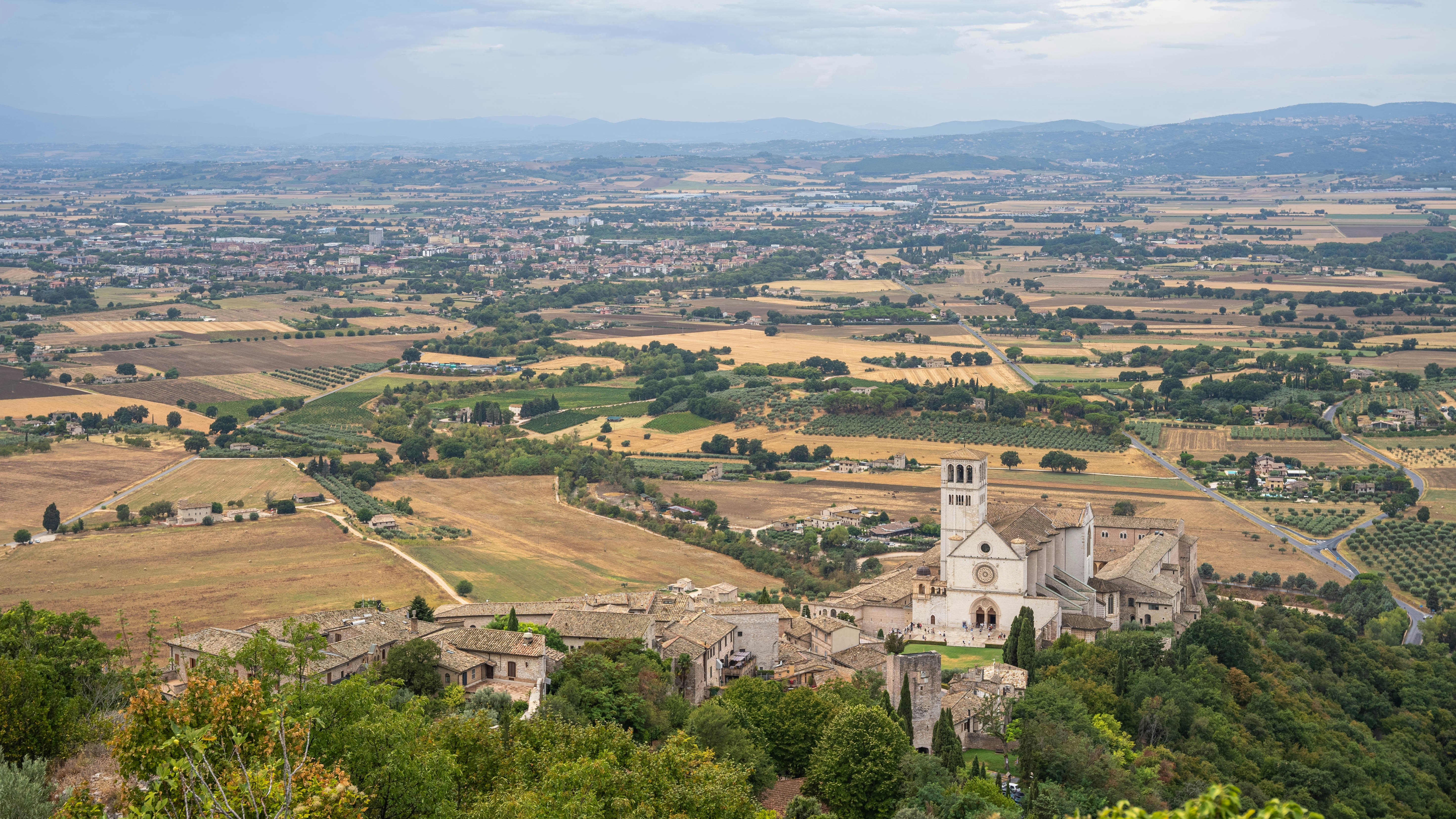 A view of a village from a hill, This scenic image captures a panoramic view of the Umbrian countryside in Italy, with the historic town of Assisi nestled in the foreground. The landscape is a patchwork of rolling fields, olive groves, and vineyards stretching out into the distance. Dominating the scene is the Basilica of St. Francis of Assisi, a significant landmark with its white stone facade and towering bell tower. The surrounding area is dotted with traditional farmhouses and winding roads, creating a picturesque and tranquil atmosphere. The distant hills and soft, overcast sky add depth to the composition.