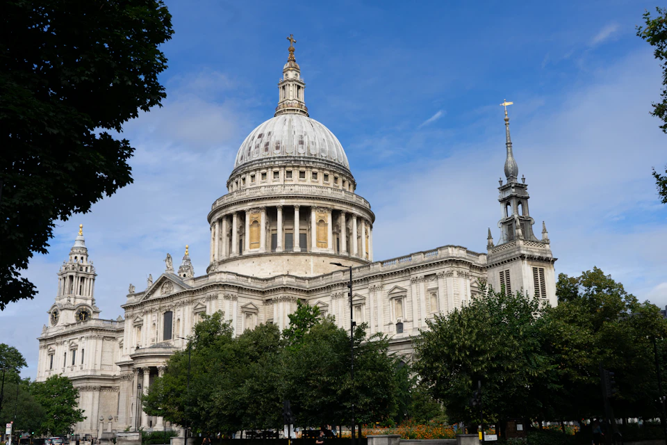 The image shows a large, white stone building with a dome and two towers on either side. The building is situated within a park-like setting, surrounded by lush green trees that provide a natural backdrop to its architectural grandeur. The perspective of the photo is from across the street, looking towards the building, which enhances its imposing presence against the sky.

The image also contains some text, but it is not clear what it says without additional context or details.