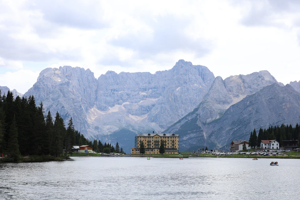 The image depicts a serene mountain landscape with a large, multi-tiered building situated on the right side. The building is a grand, two-story structure with multiple levels and balconies, suggesting it may be a hotel or resort. In front of the building, there's a lake that reflects the surrounding scenery, adding to the picturesque nature of the scene. Several boats are docked at the shoreline, indicating recreational activities taking place in this area. The mountains in the background provide a stunning backdrop for the lake and the building, creating an overall sense of tranquility and natural beauty.