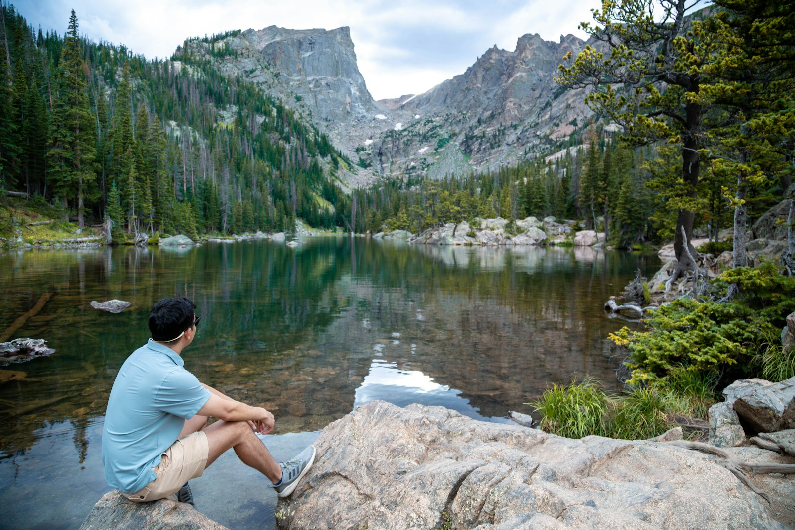 A hiker sitting on a large boulder at the edge of a clear green alpine lake beneath high mountain walls