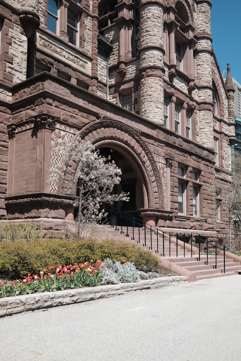 The image shows a large, ornate stone building with multiple levels and arched windows. The building is situated on a street corner, surrounded by lush green bushes and trees. A sidewalk runs along the front of the building, leading up to an entrance that features a black railing. In front of the building, there are red flowers blooming in a well-maintained garden with various plants and shrubs.