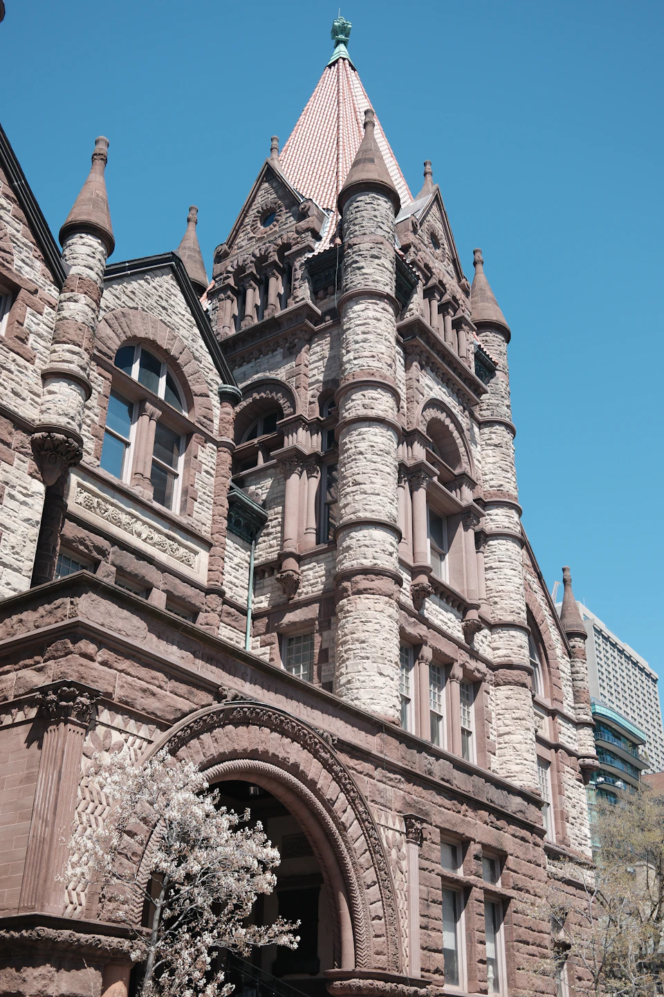The image shows a large, ornate stone building with multiple levels and towers reaching towards the sky. The building is bathed in sunlight, highlighting its intricate architectural details. A tree can be seen on the left side of the frame, adding a touch of nature to the man-made structure.

The perspective of the photo is from below, looking up at the imposing height of the building and the tower it sits atop. The image does not contain any discernible text or countable objects, making the scene appear serene and majestic.