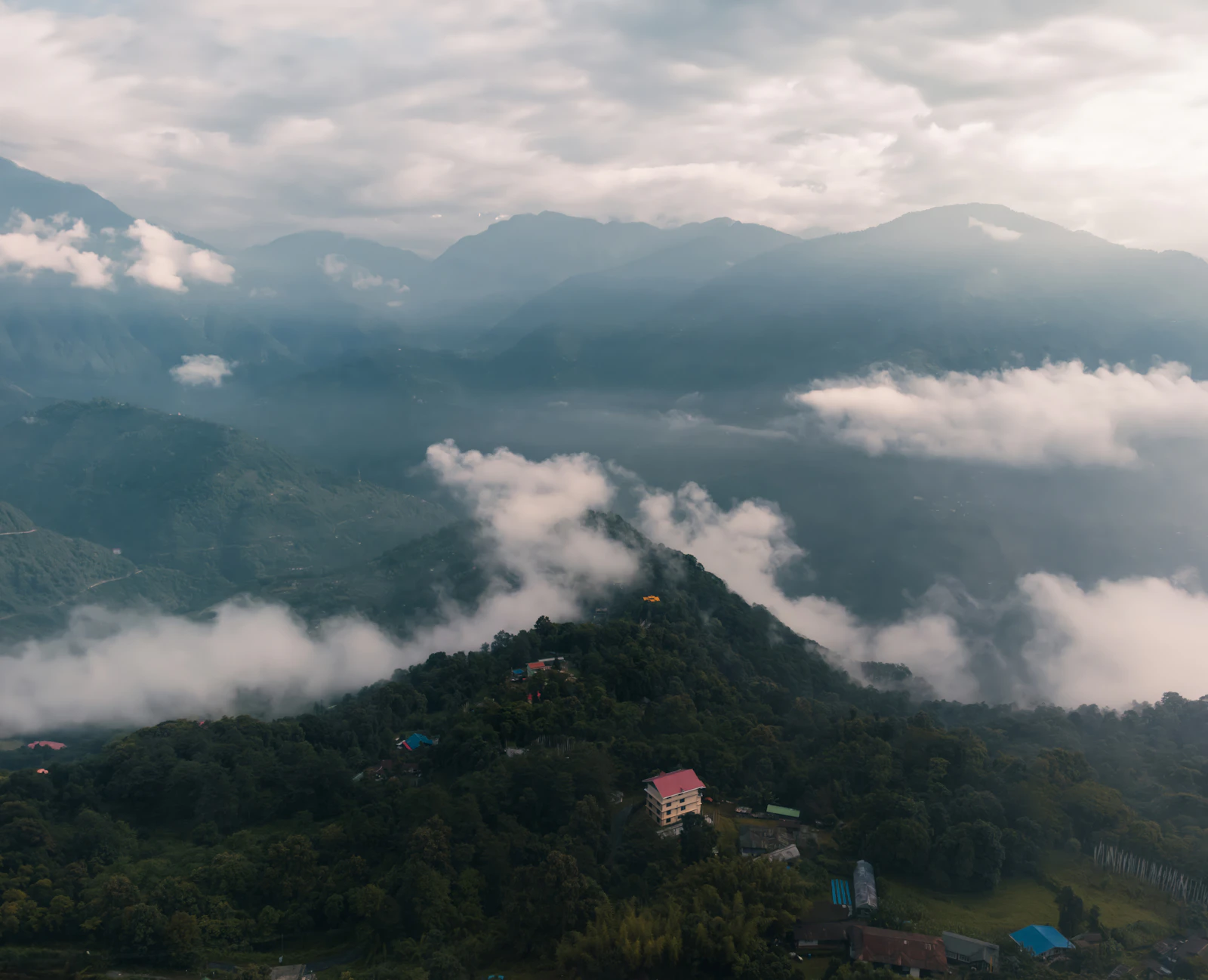 Cloud pooling in the valleys below the Pelling ridge in West Sikkim, houses scattered over its forested slope under a grey sky