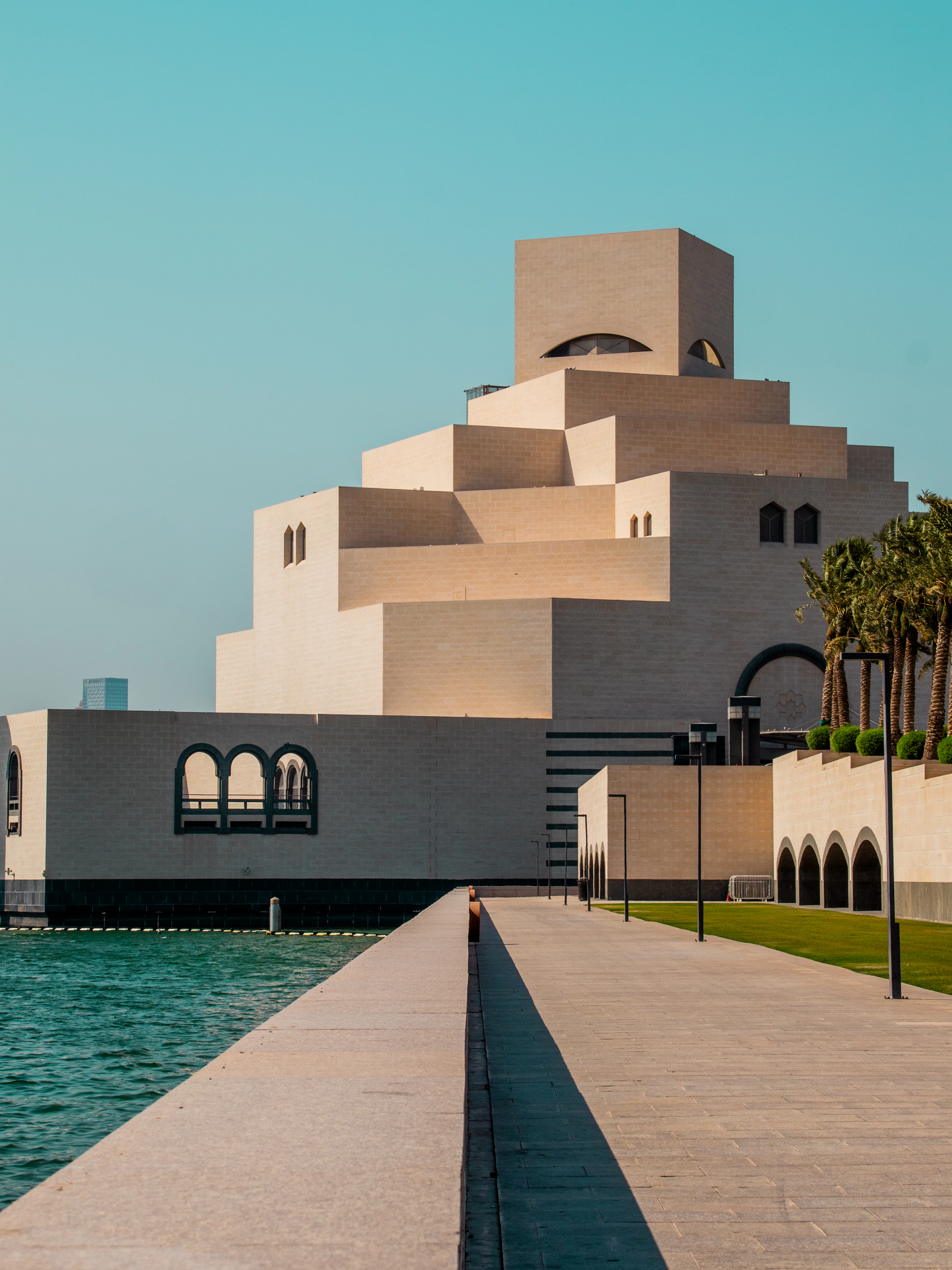 I. M. Pei's Museum of Islamic Art in Doha at golden hour, its stacked cream limestone blocks and the shadowed oculus of its upper tower glowing warm above a long empty causeway and the turquoise water of the bay