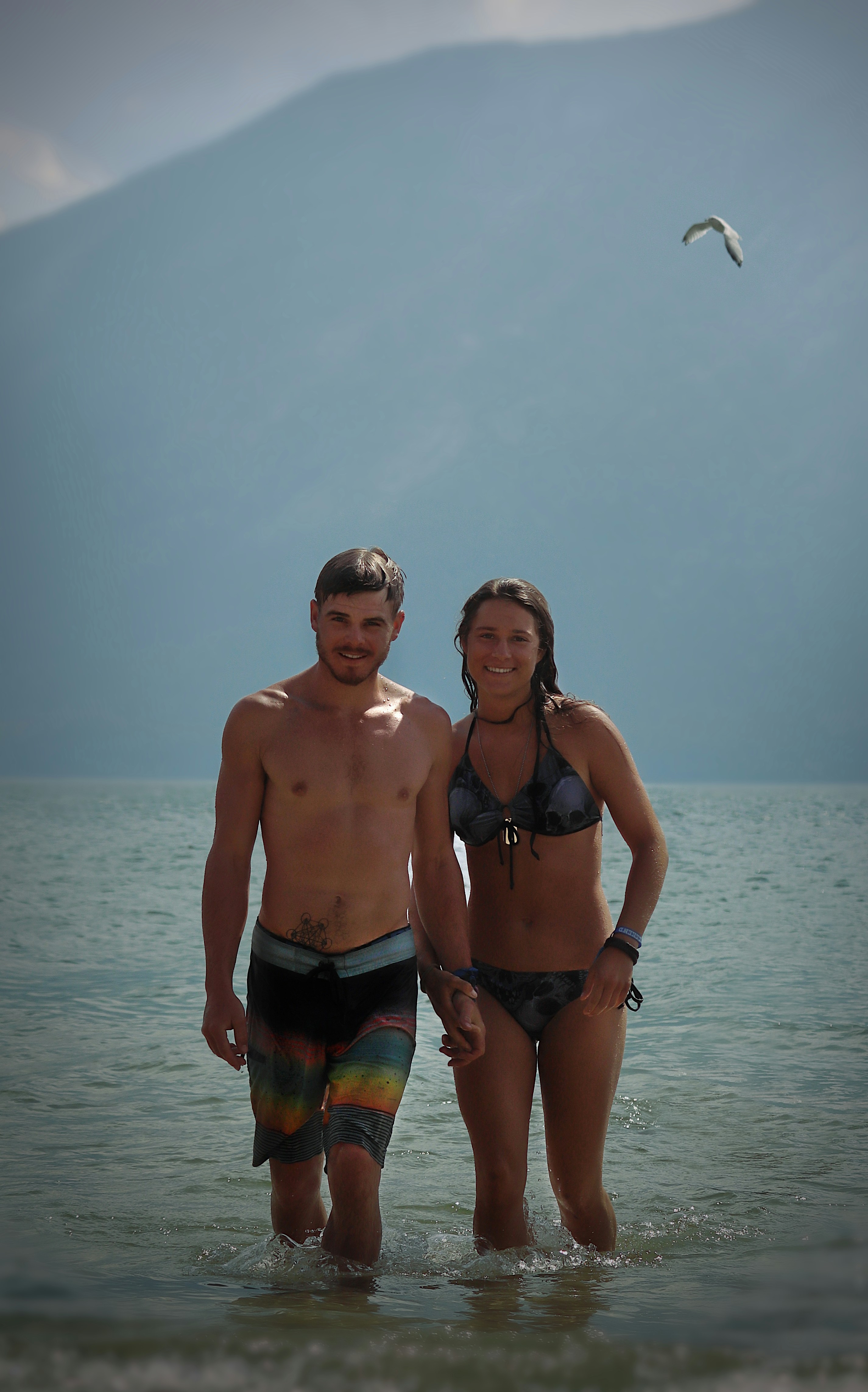 Couple walking hand-in-hand through shallow water at the beach, enjoying a sunny day, with mountains in the background.