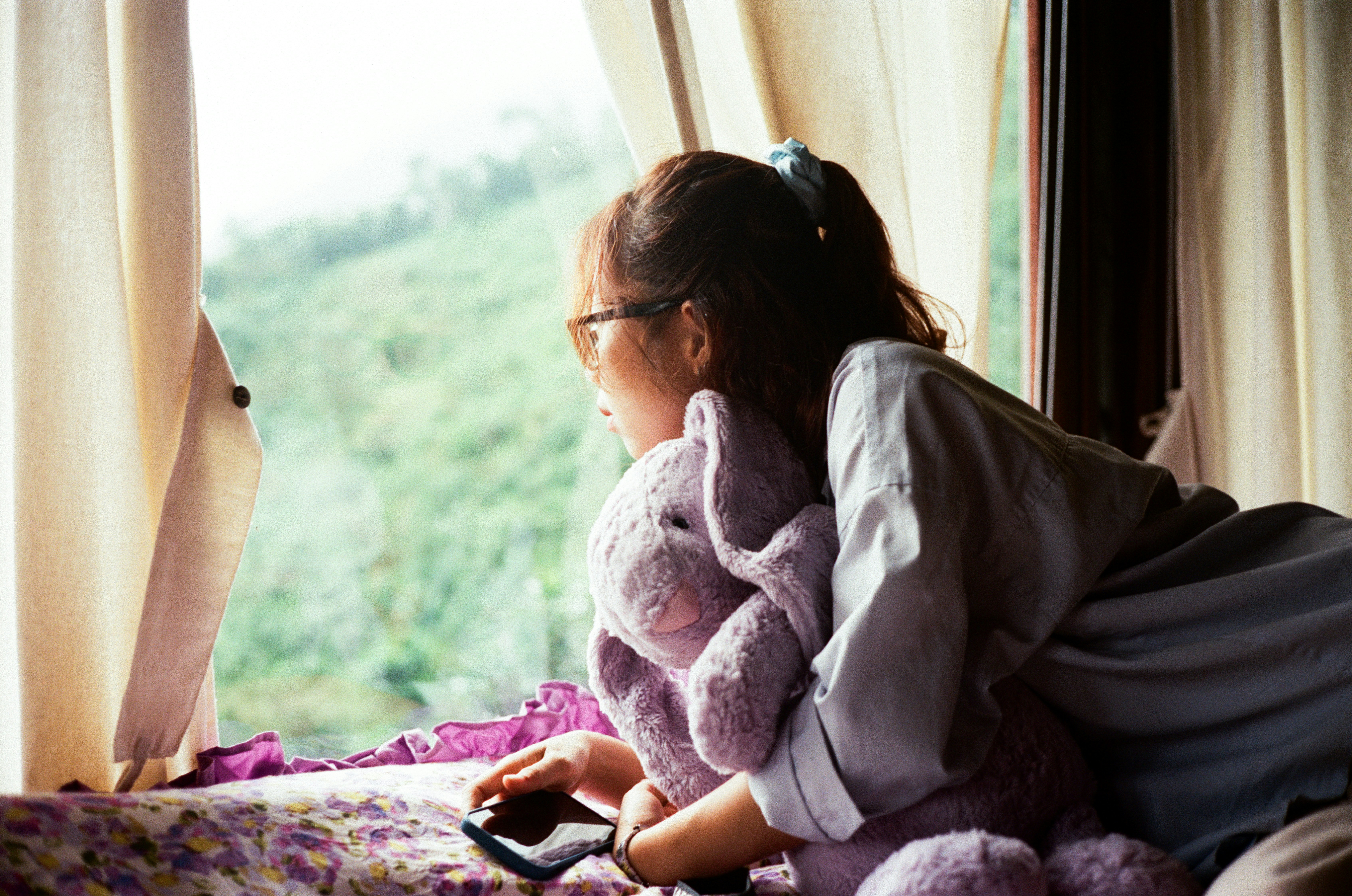 Photo of a person sitting on their bed without a phone, looking out of a window with morning light, wearing casual home clothes.